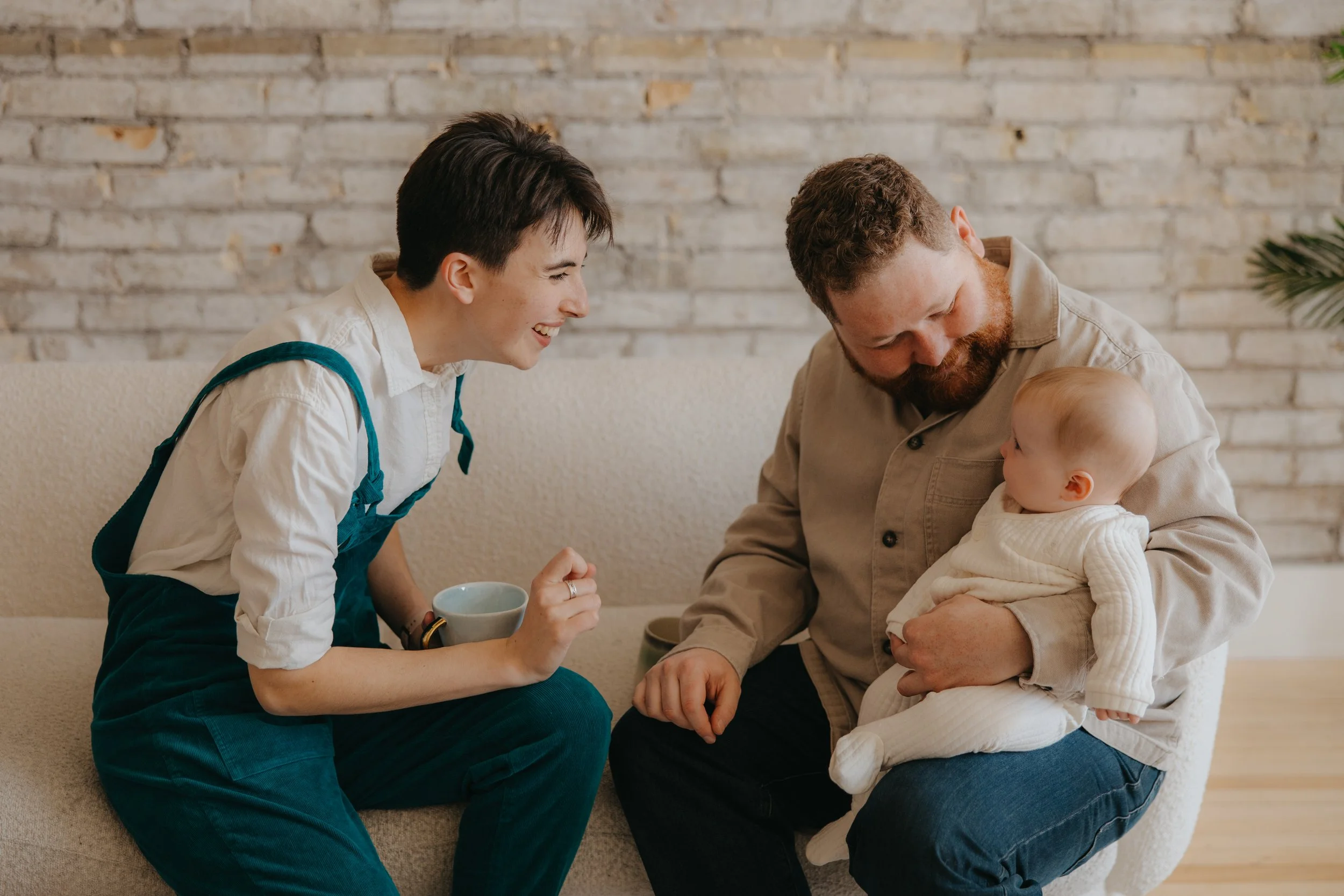 A woman laughing while sitting on a beige couch, holding a mug, engaging with a man and a baby. The man is sitting on a chair, holding a baby girl dressed in white. They are indoors with a brick wall in the background.