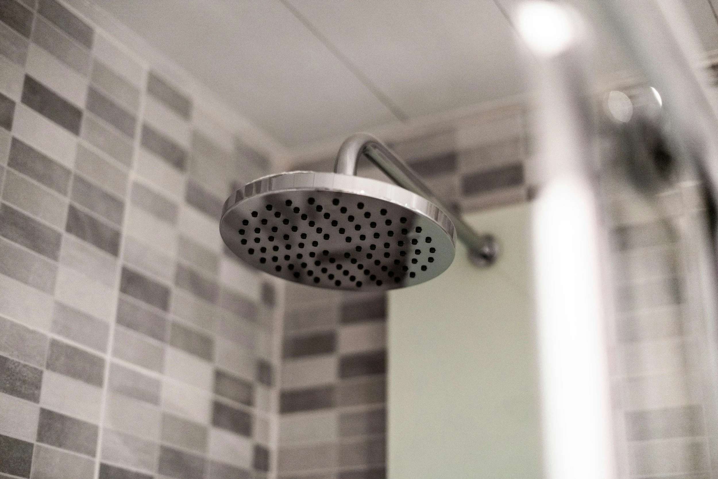 Close-up of a modern, round, silver showerhead mounted on a wall in a bathroom with gray and white tile.