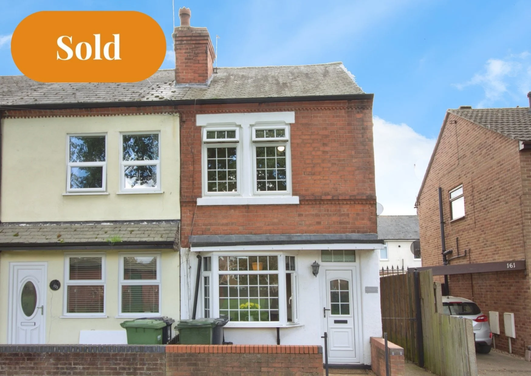 A three-story house with a combination of brick and cream-colored exterior, sold sign on the top left corner, a white front door, a large bay window with a white frame on the ground floor, and two double-hung windows on the upper floor. There are green trash bins in front and a small brick wall along the front yard.