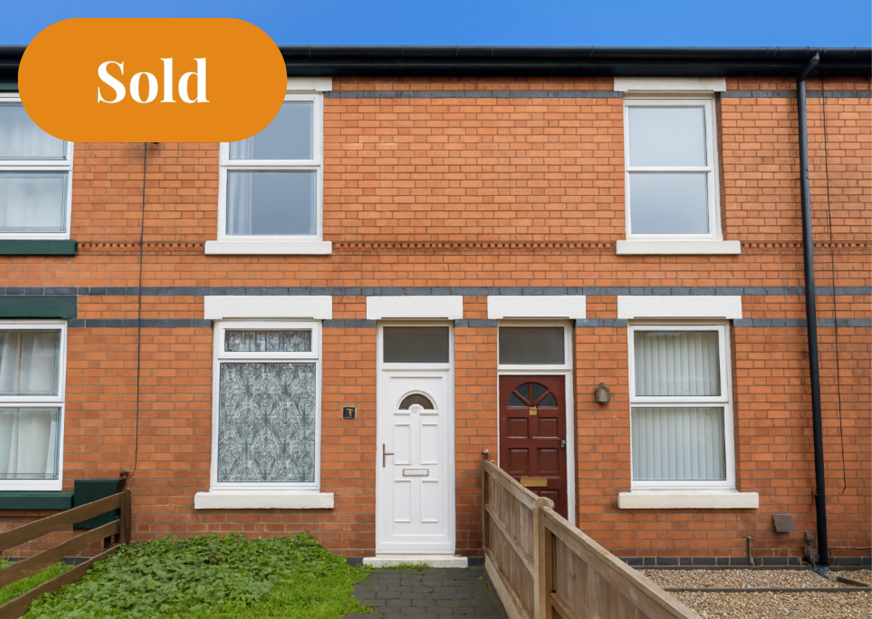 A brick townhouse with white window frames, a white front door, and a red door, with a small garden and fencing in front. A large orange "Sold" sign is in the upper left corner.