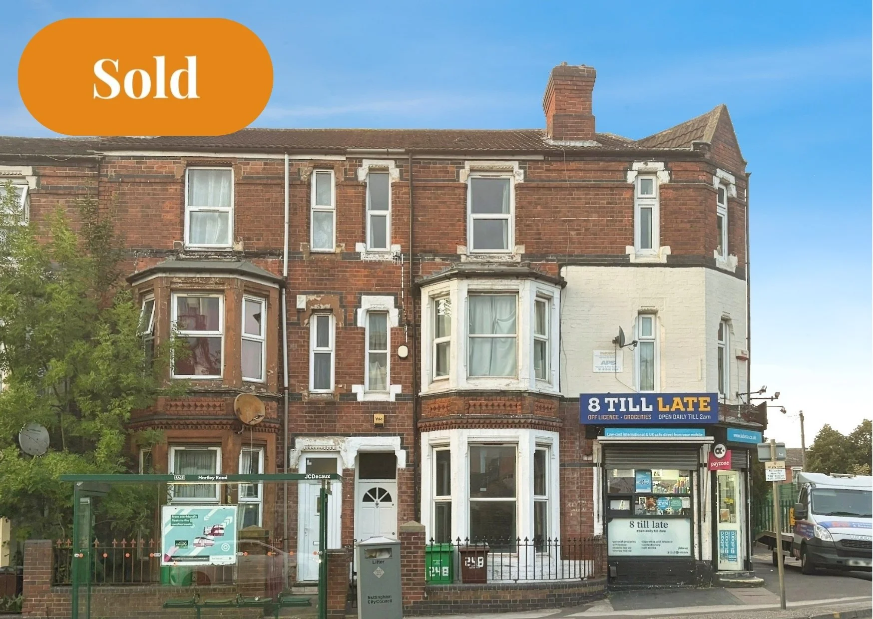 A three-story brick building with some white trim and bay windows, with a sign that reads '8 TILL LATE' above a small storefront on the ground floor. There is a bus stop in front labeled 'Hartley Road', a trash bin, and a parking vehicle to the right. A tree with green leaves is on the left side of the image.