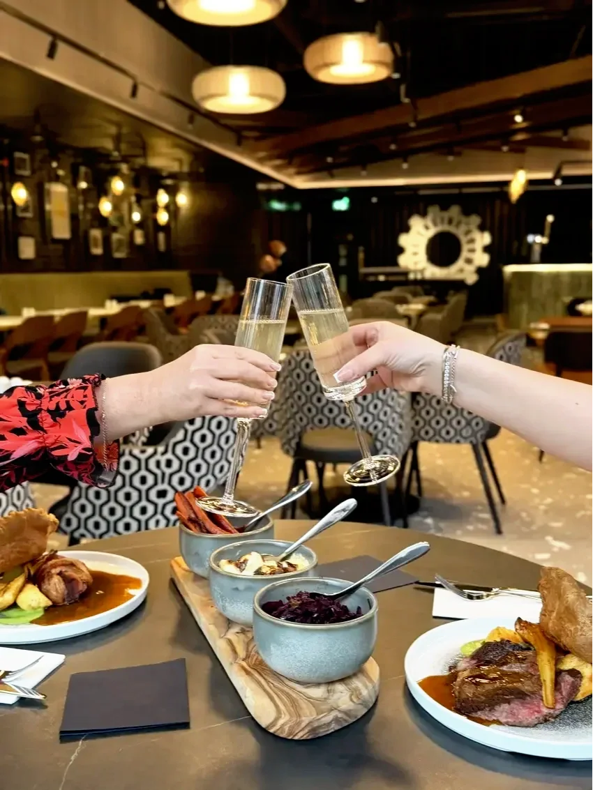 Two people clinking champagne glasses at a dining table with plates of food and bowls of condiments in a cozy restaurant setting.