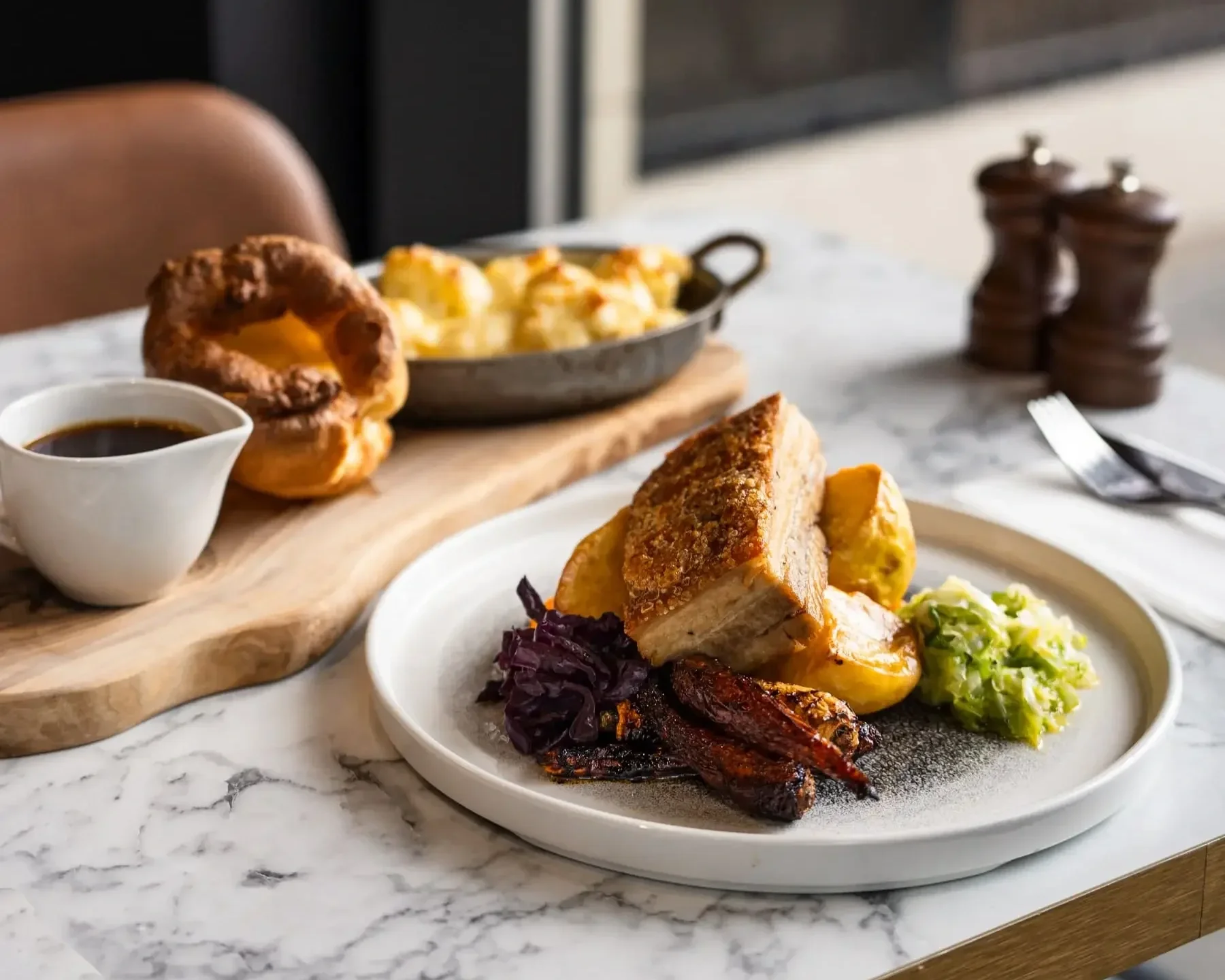 Roast pork loin with elevated sides on a white ceramic plate. Alongside is a serving platter with gravy, Yorkshire pudding and a tray of cauliflower cheese, on at table at The Engine Room in Derby