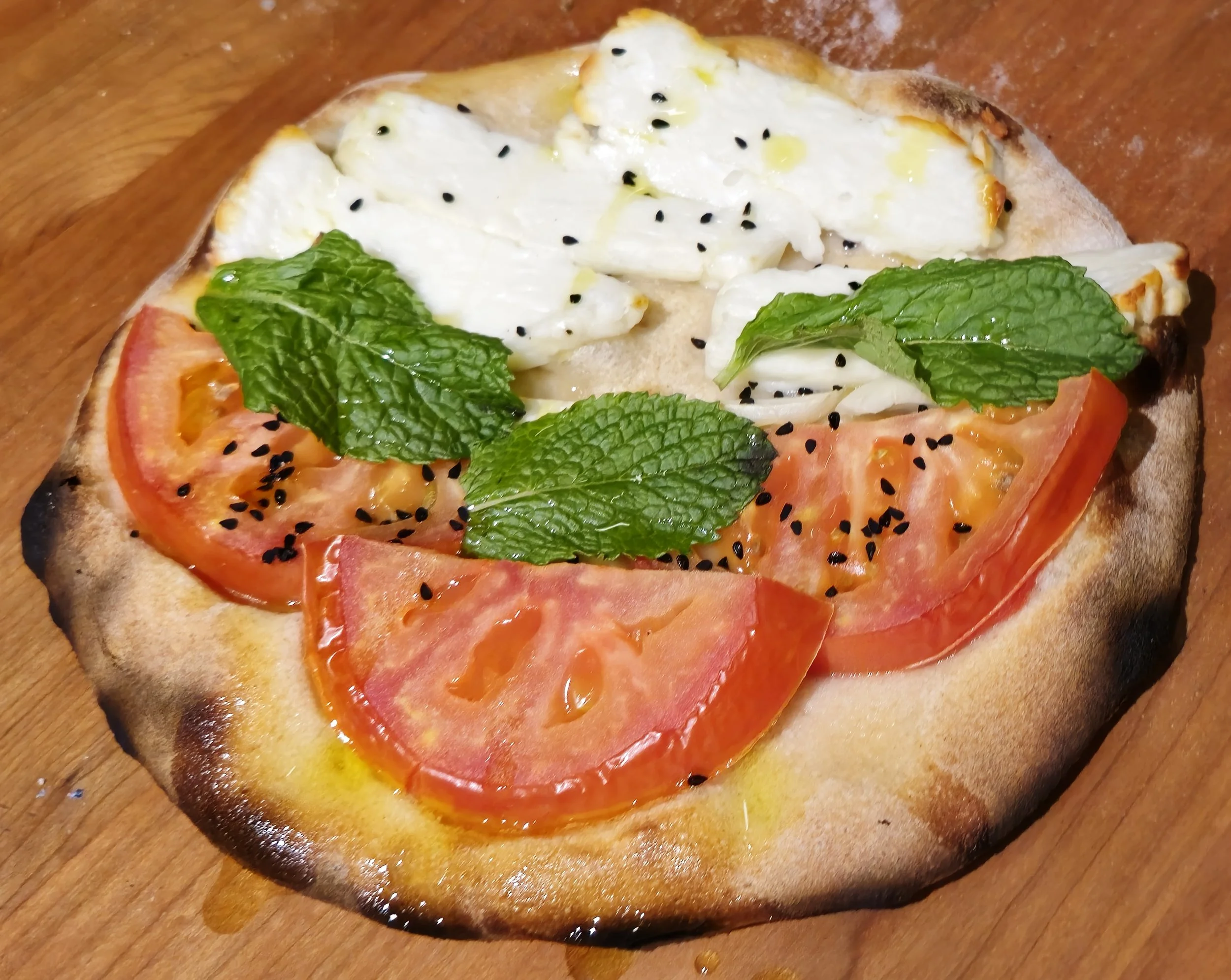 Close-up of a pizza with tomato slices, fresh basil leaves, mozzarella cheese, and black sesame seeds on a wooden surface.
