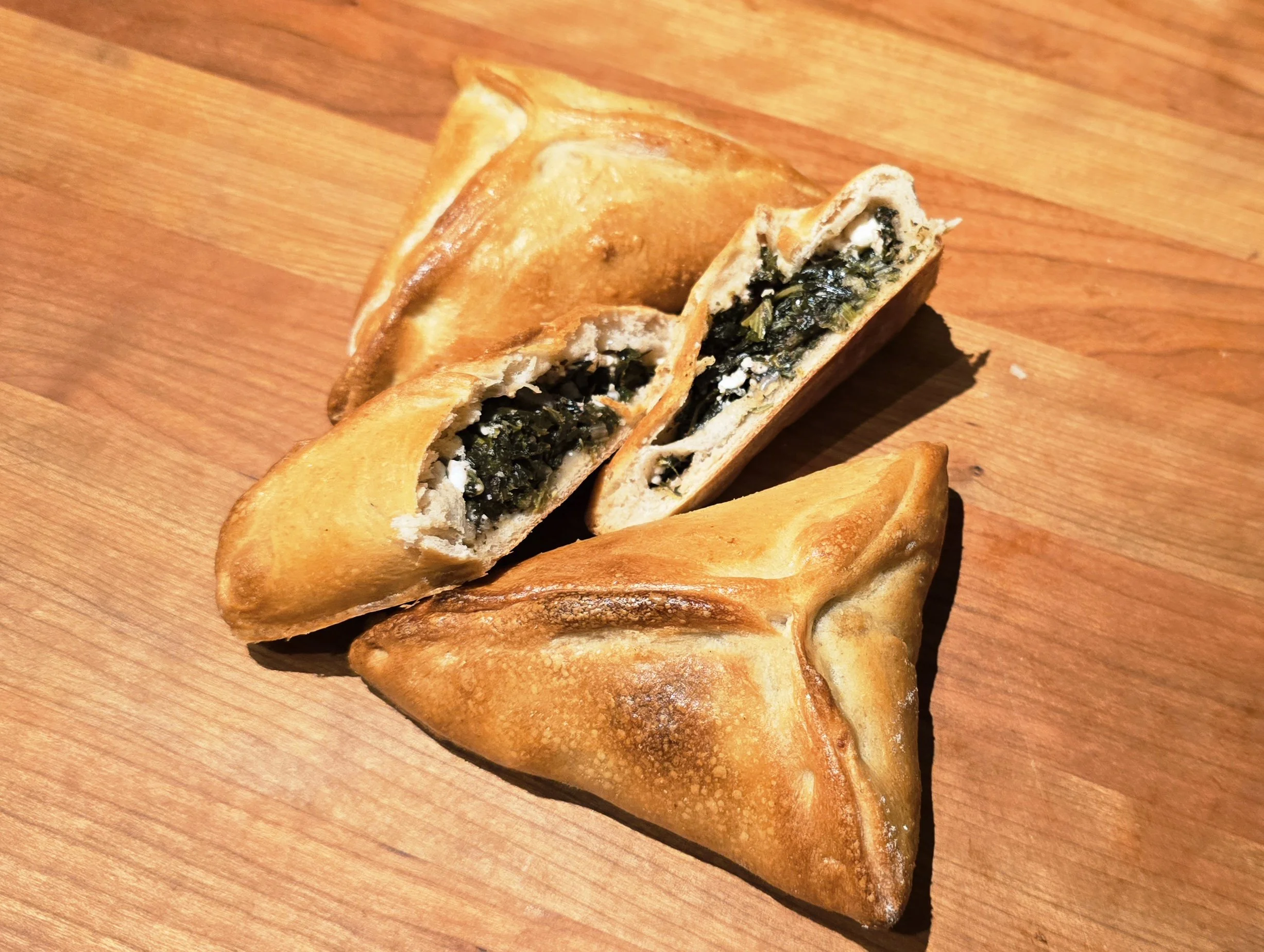 Three golden-brown fried pastries, one of which is cut open to reveal a dark leafy vegetable filling, on a wooden surface.