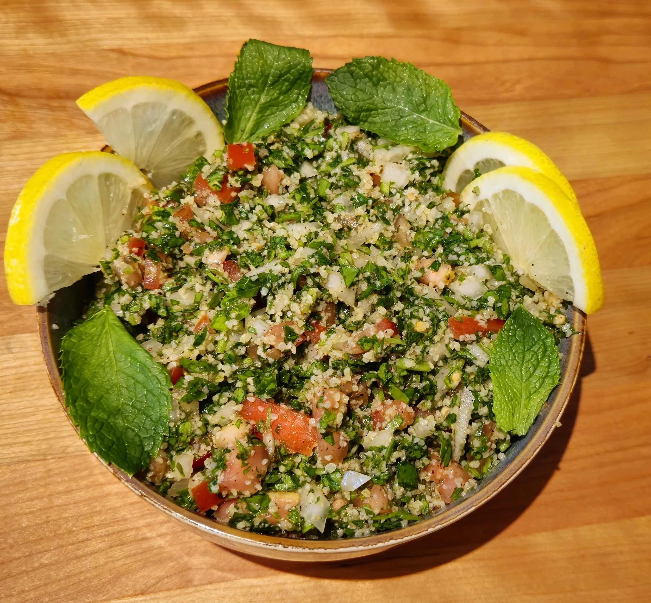 Bowl of tabbouleh salad garnished with lemon wedges and mint leaves, on a wooden surface.