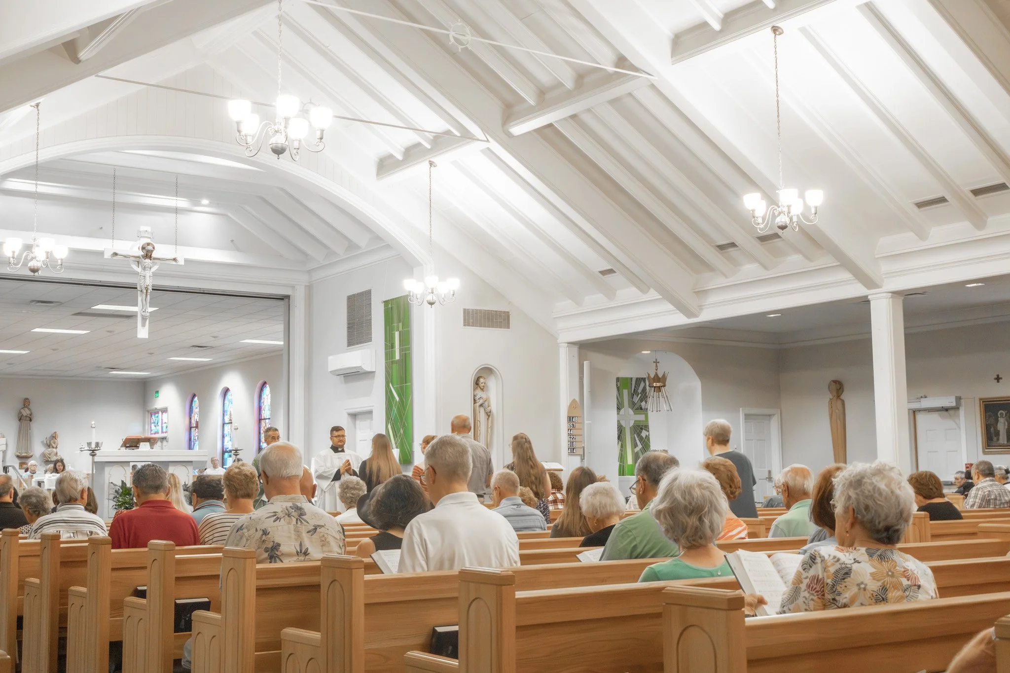 Inside St. Albert the Great Catholic Church in Kettering, Ohio, during a service with people seated in pews, priests at the altar, stained glass windows, religious statues, and chandeliers hanging from the ceiling.