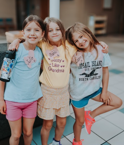 Three young girls standing together, hugging, smiling, in an indoor setting.