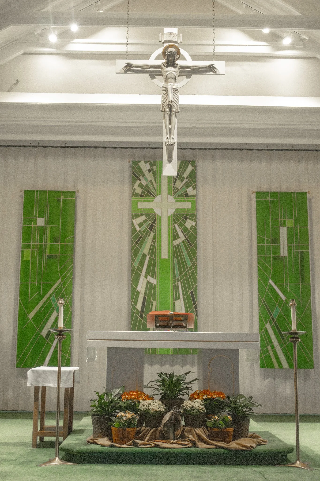 Interior of a church altar with a white crucifix above, green and white geometric banners, and flowers arranged on the altar table.