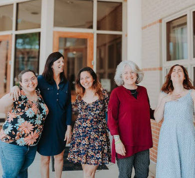 Five women standing outside a building, smiling and laughing together.