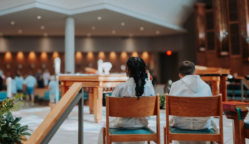 Two children sitting in church, facing the altar, with a blurred congregation in the background.