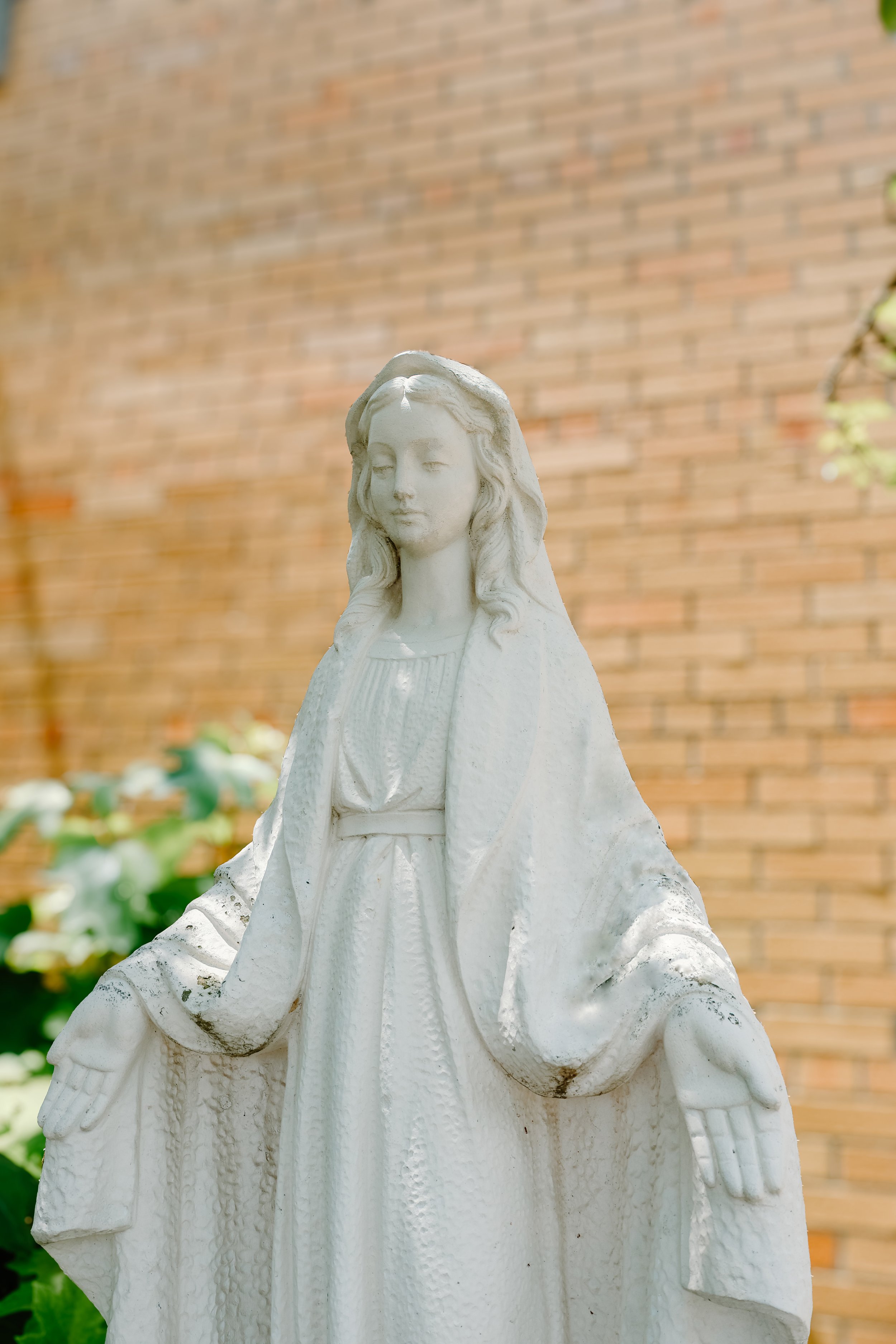 White statue of Jesus Christ with a serene expression, standing outdoors against a brick wall, with greenery in the background.
