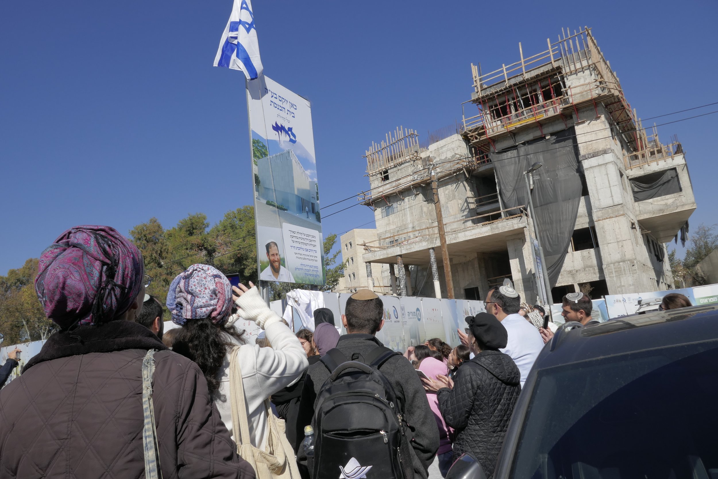 Crowd gathered near construction site with building framework and a sign featuring Rav Avi.