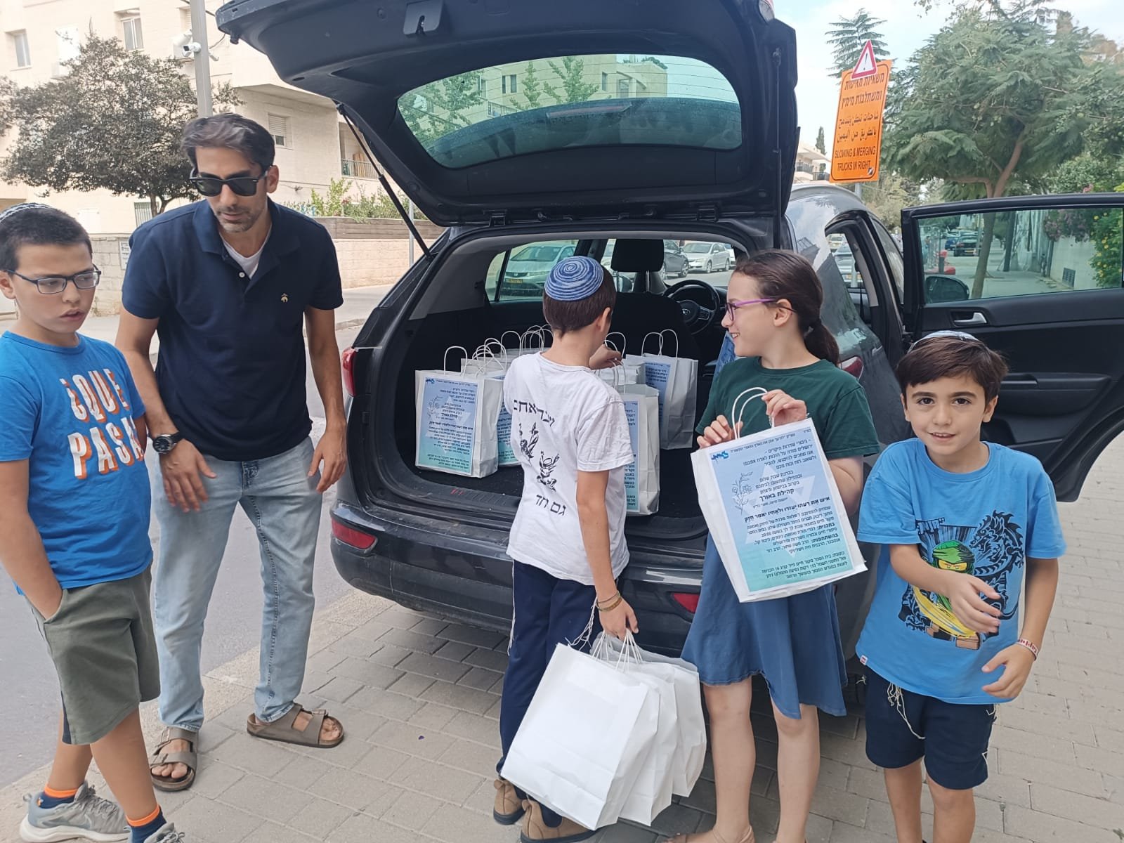 Group of kids and an adult unloading bags from a car trunk on a street.