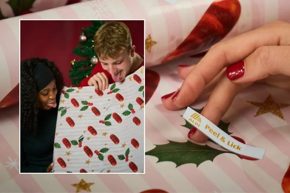 Close-up of a hand with red nail polish peeling a sticker labeled 'ALDI Peel & Lick' from a Christmas gift wrapped in holiday-themed paper. In the background, two people joyfully have fun with a large wrapped gift, with Christmas tree visible.