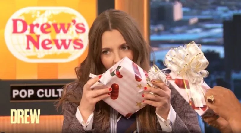 A woman with long brown hair is holding a Christmas gift wrapped in holiday paper with cherry and star designs, and another gift with a bow in front of her face, during a TV show set with a Drew's News logo in the background.