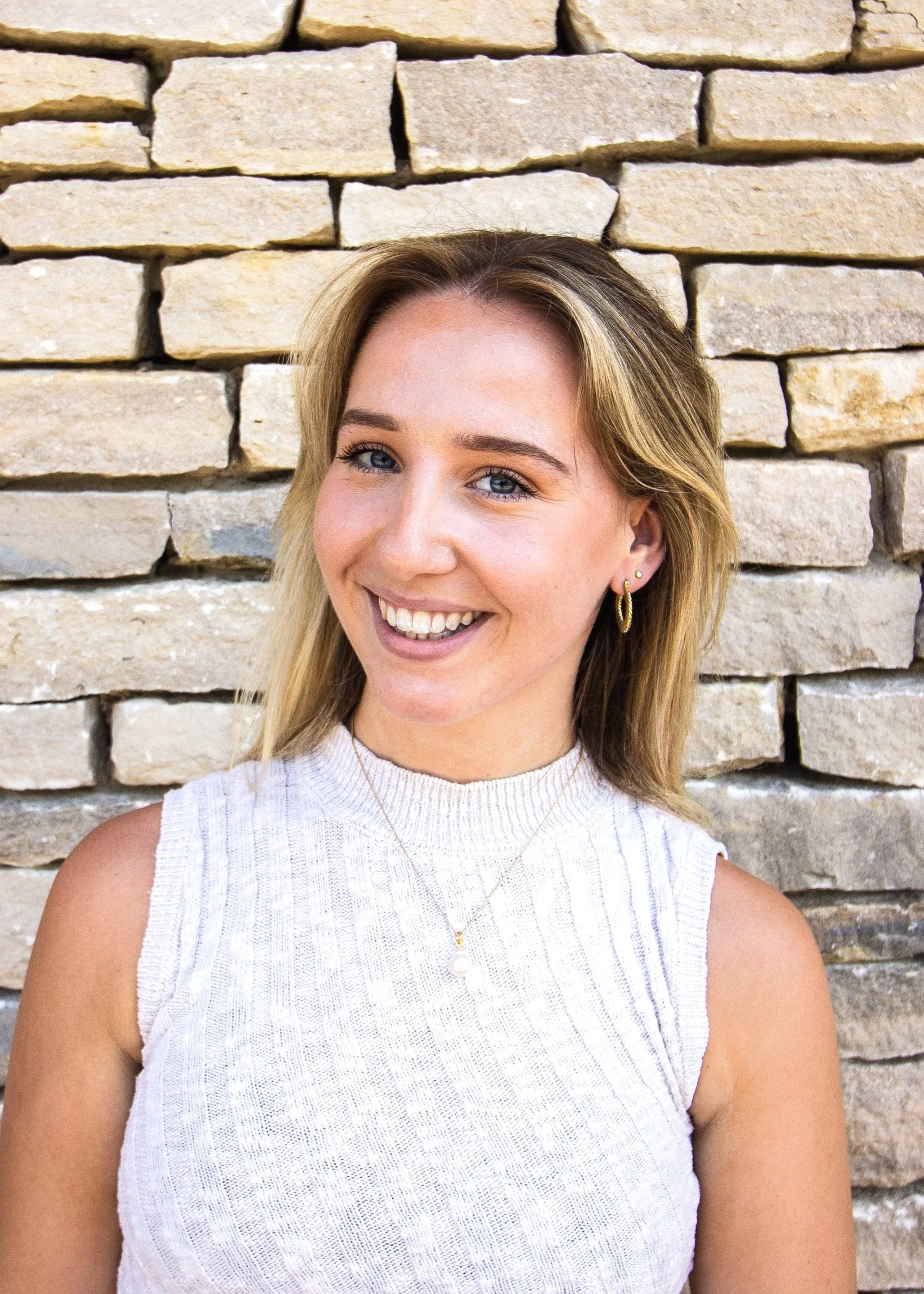 Smiling person standing in front of a brick wall, wearing a sleeveless white sweater and a necklace.