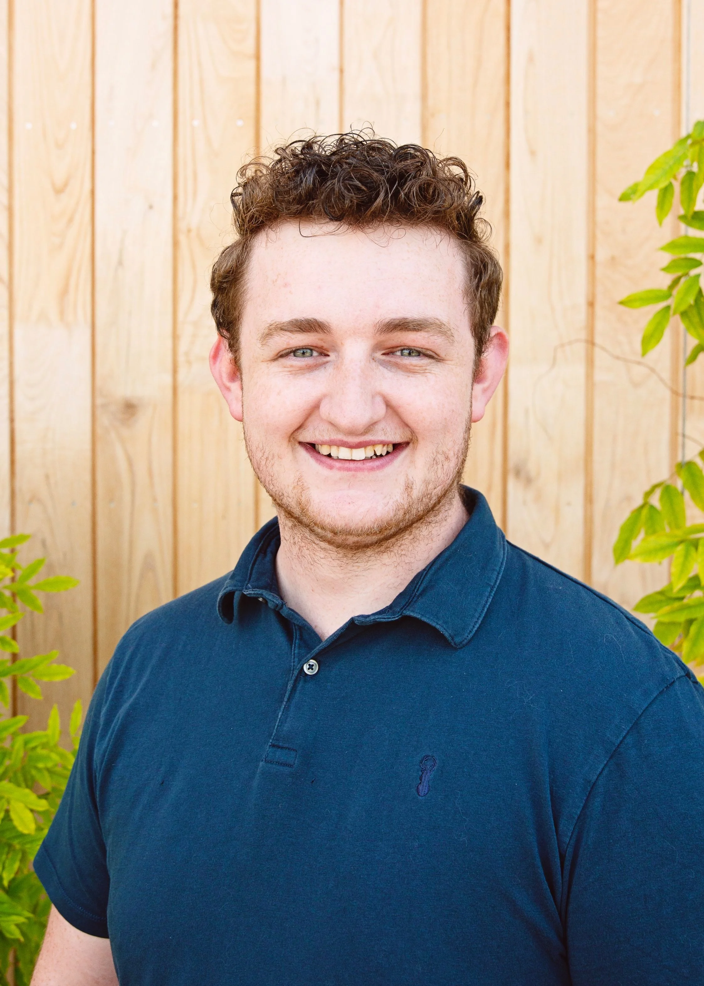 Smiling man wearing a navy polo shirt, standing in front of a wooden wall with green foliage on the side.