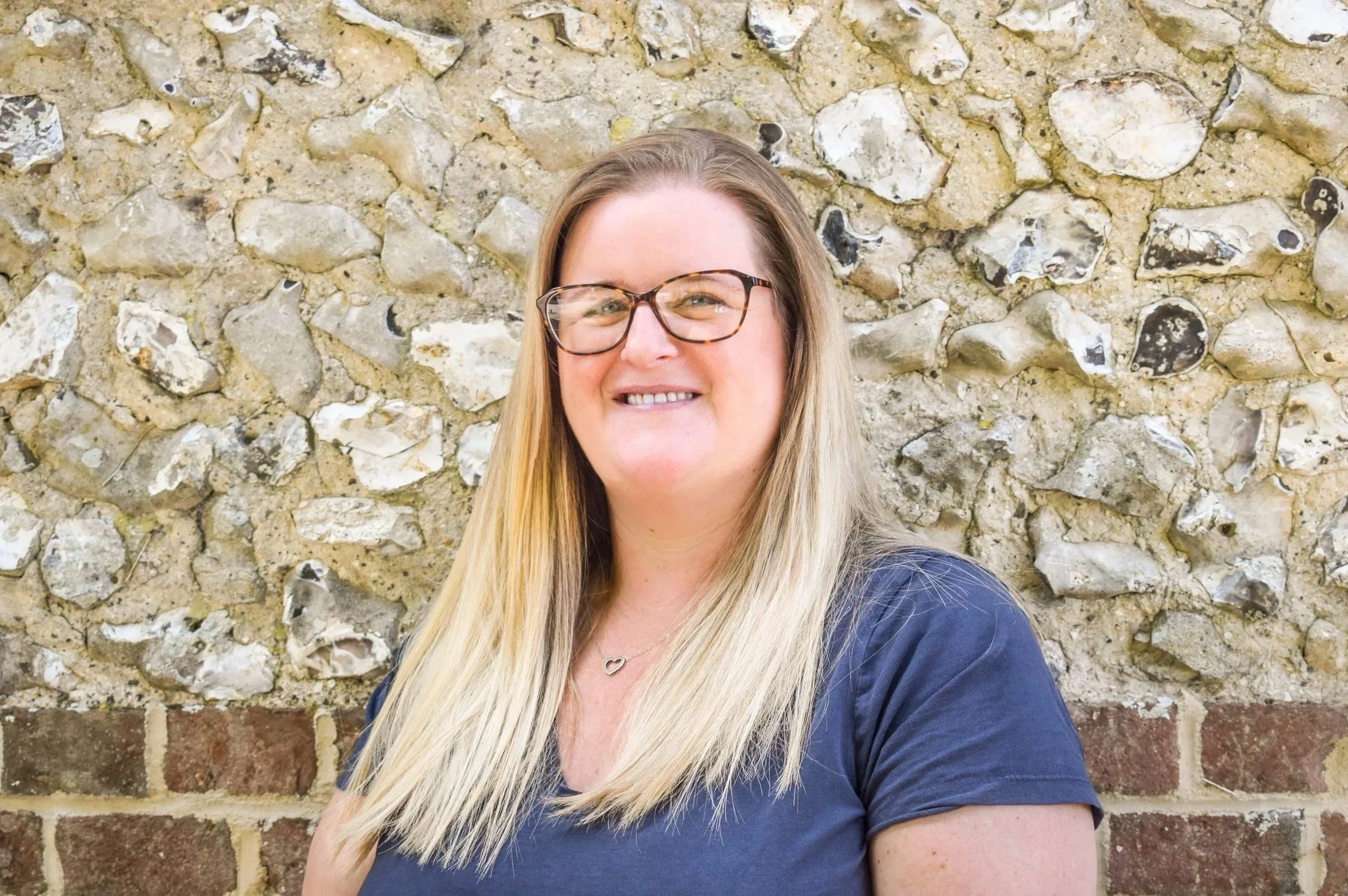 Hannah Fraser, account lead, smiling at the camera, standing in front of a stone and brick wall, wearing glasses and a navy blue t-shirt.