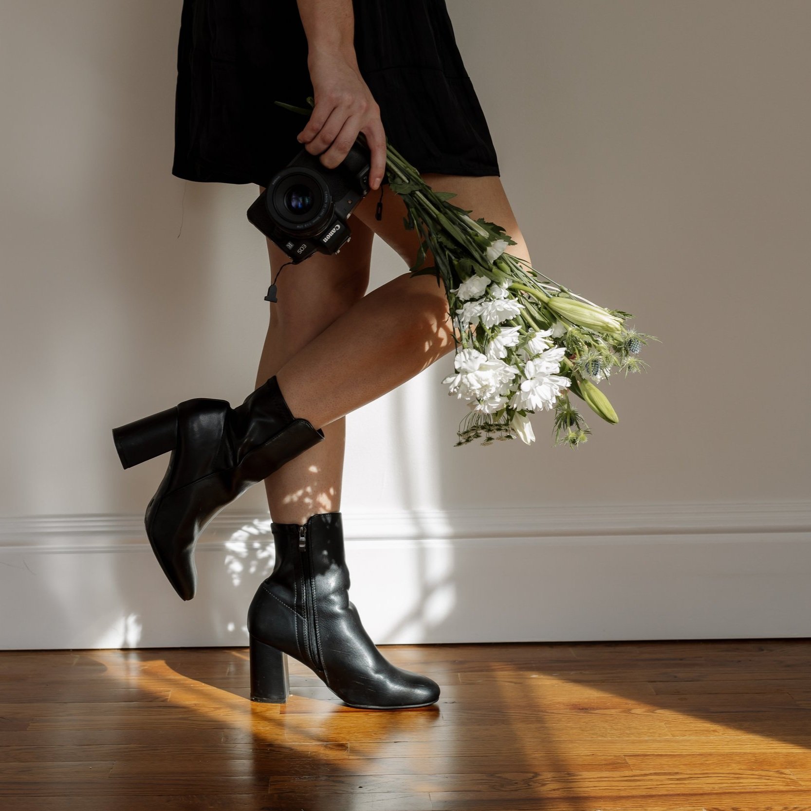 Person wearing black boots and a black dress holding a camera in one hand and a bouquet of white flowers behind their legs, standing against a plain wall.
