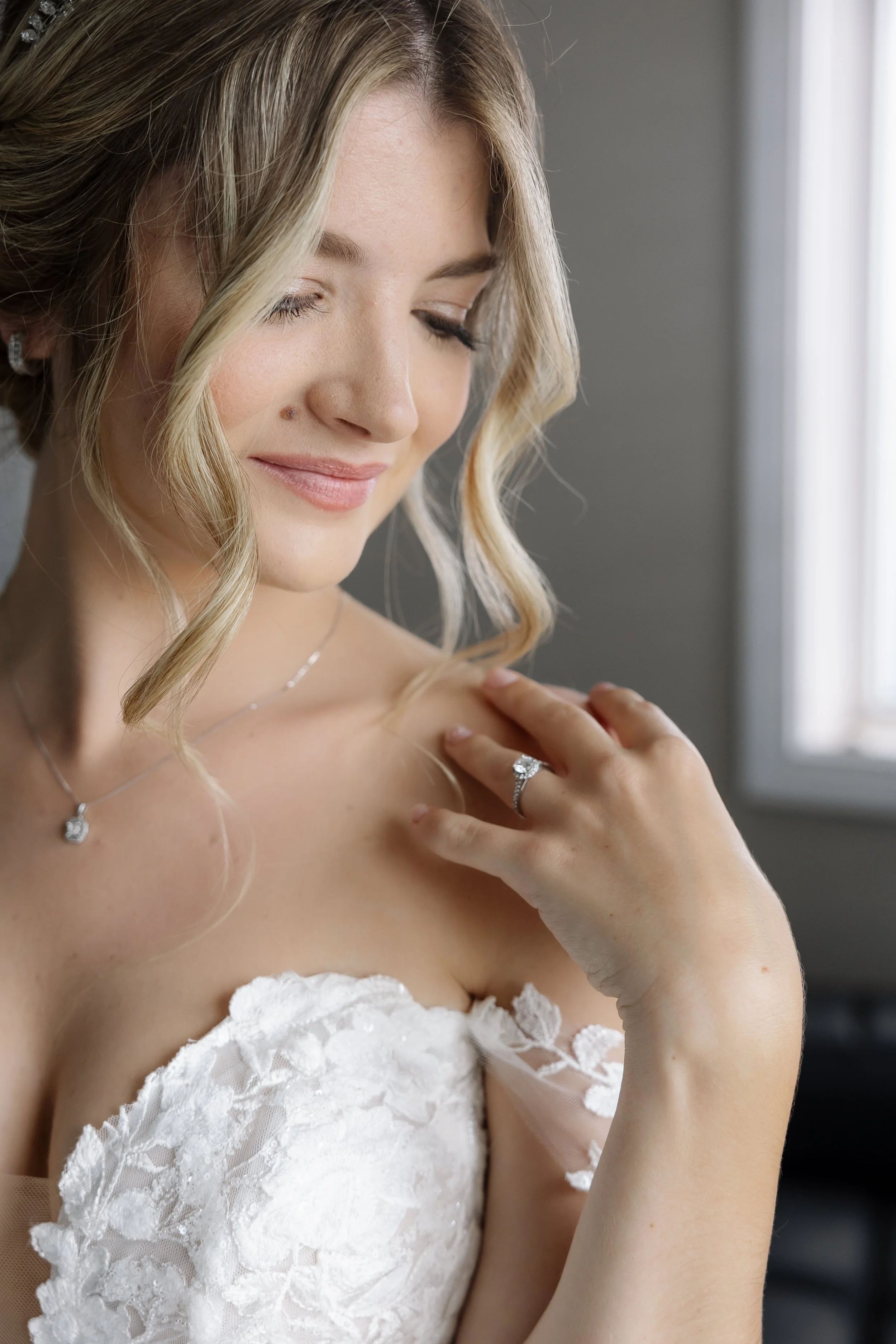 A bride with blonde hair styled in loose curls, wearing a white lace strapless wedding dress and jewelry, gently touching her shoulder with a serene expression.