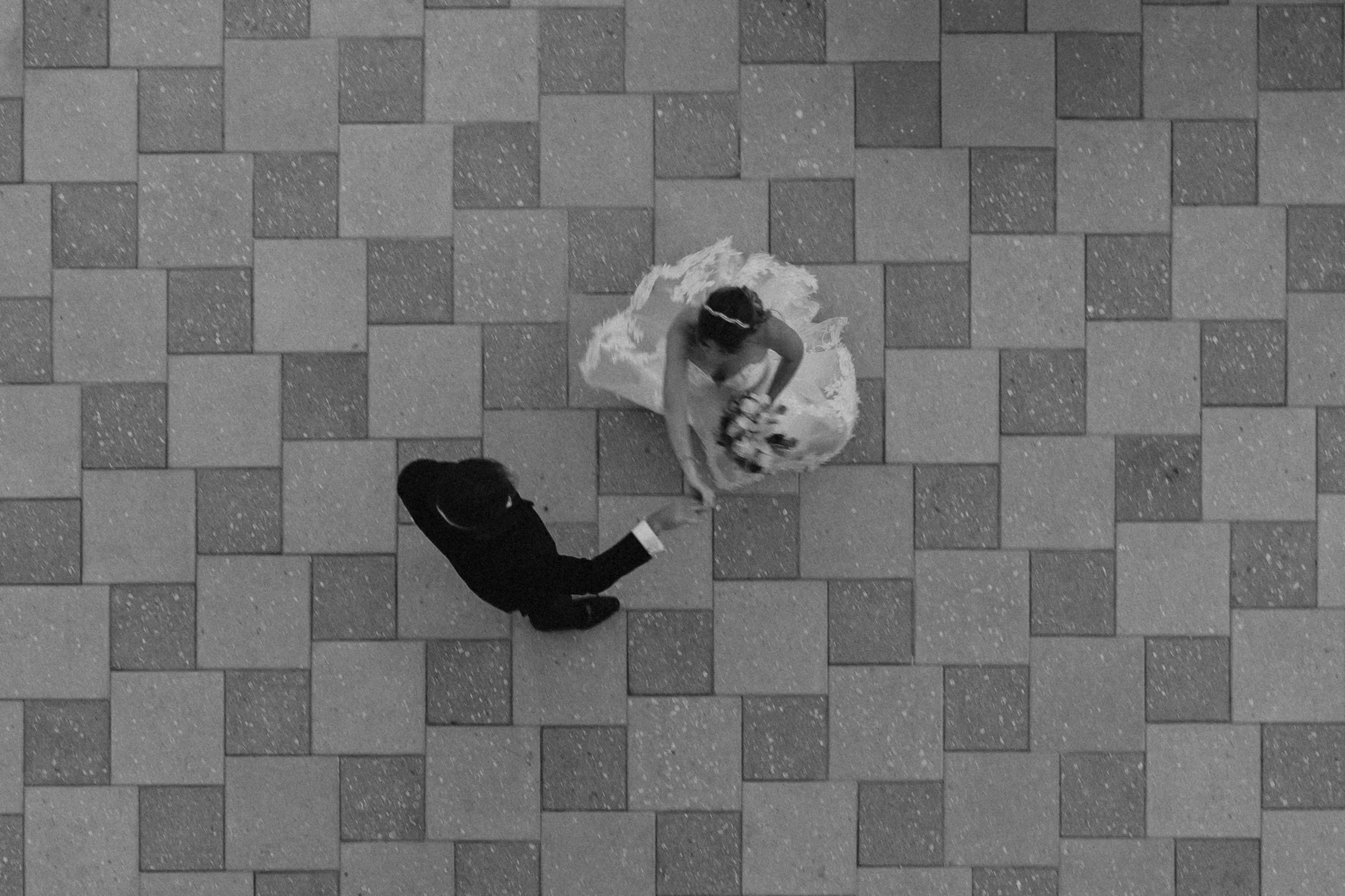 A bride in a wedding dress and a groom in a suit are dancing on a tiled floor. The bride is holding a bouquet, and the groom is reaching out to her.
