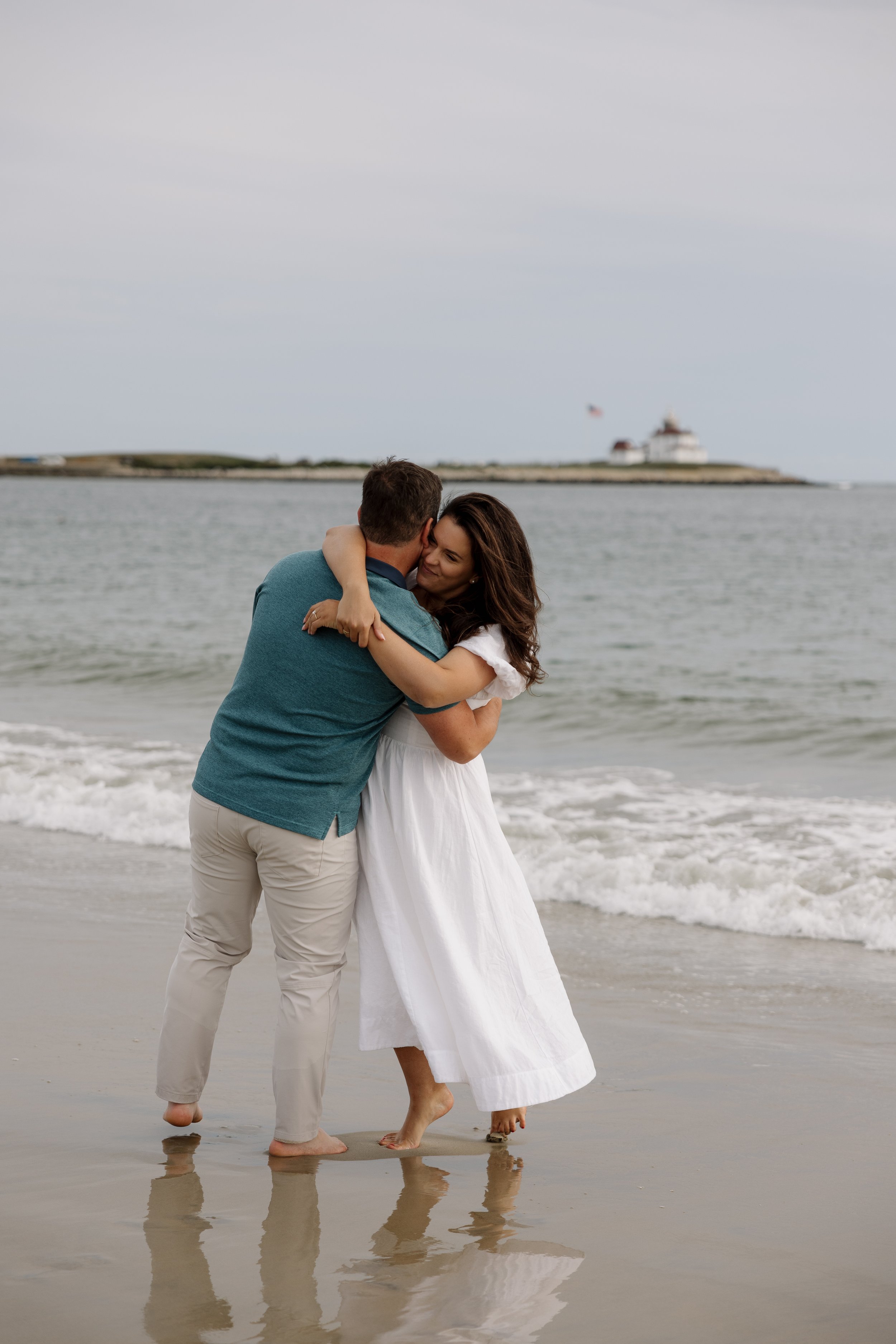 A couple embracing on a beach, standing in shallow water with waves, with an island and building with a flag in the background.