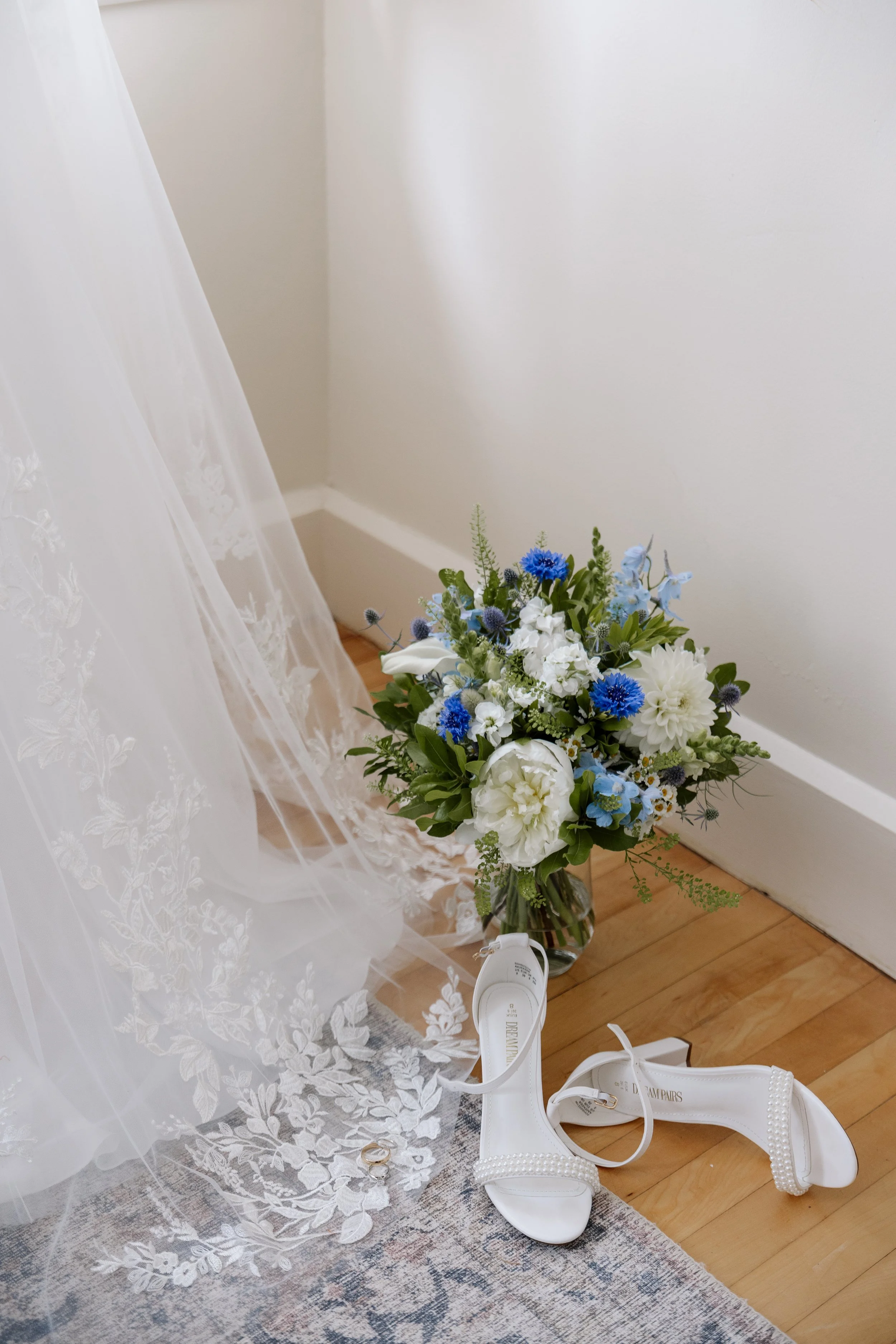 White bridal veil, a bouquet of white and blue flowers, and white high-heeled shoes with pearl embellishments on a wooden floor.