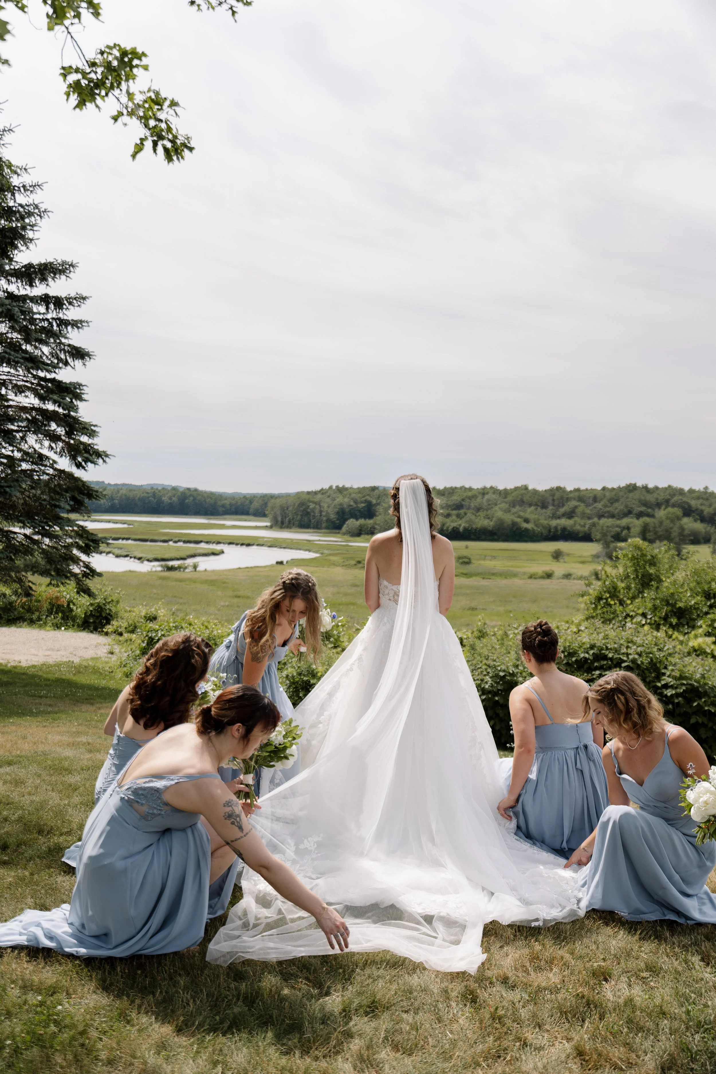 Bride in a white wedding gown and veil standing on a field with six bridesmaids in light blue dresses, kneeling around her with flowers, outdoors with a view of green fields and water in the background.