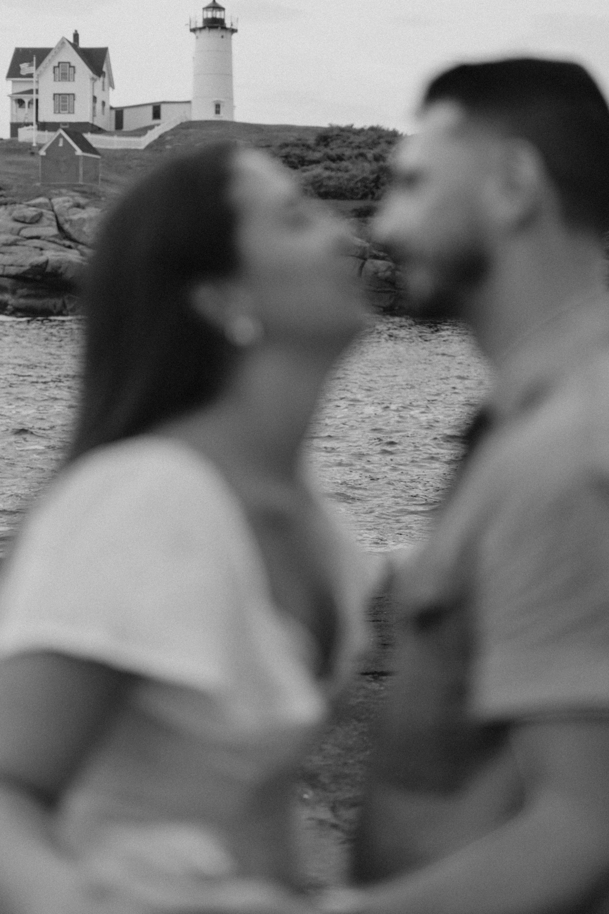 A blurred black-and-white photo of a couple kissing near water with a lighthouse and houses in the background.