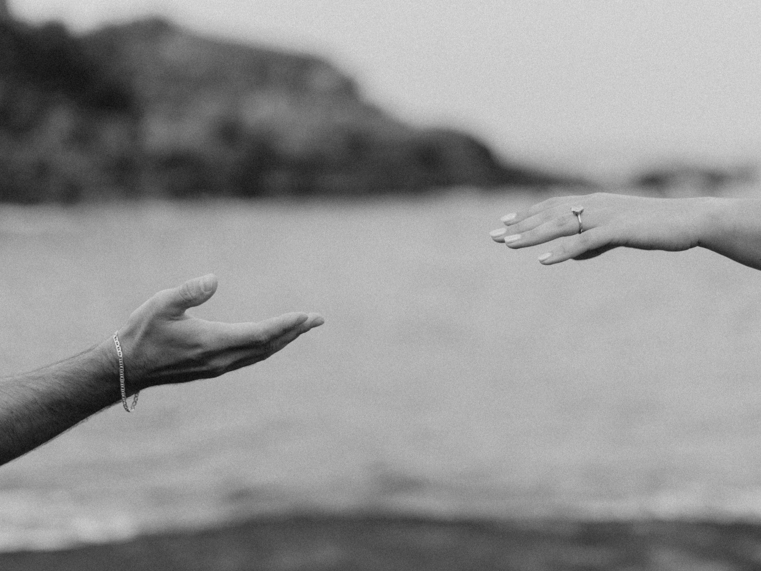 Two hands reaching towards each other over the ocean with a beach and distant cliffs in the background, black and white photo.