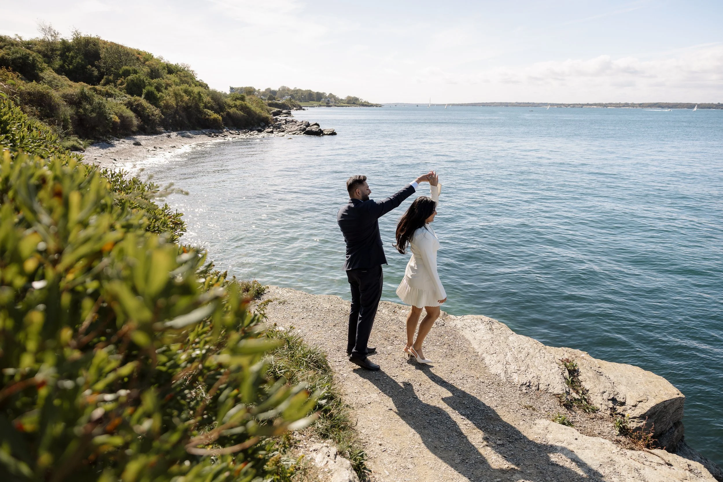 A couple in formal attire standing on a rocky shoreline, with the man holding the woman's hand above her head as they pose for a photo against a scenic waterside background.