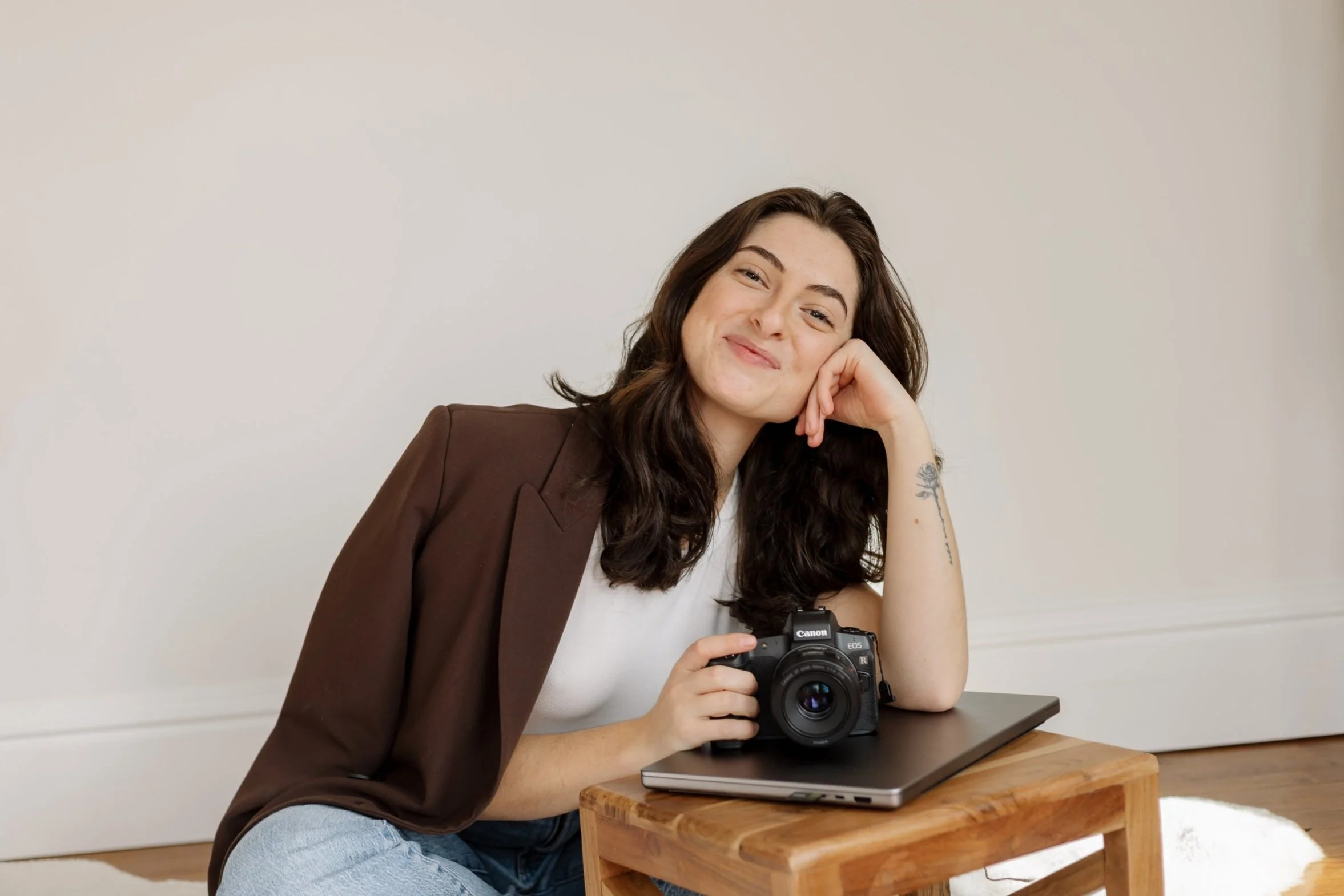 A young woman with dark hair, wearing a brown blazer and white shirt, sitting on the floor with a camera in her hand, smiling at the camera, with a laptop and a closed book on a small wooden table beside her.