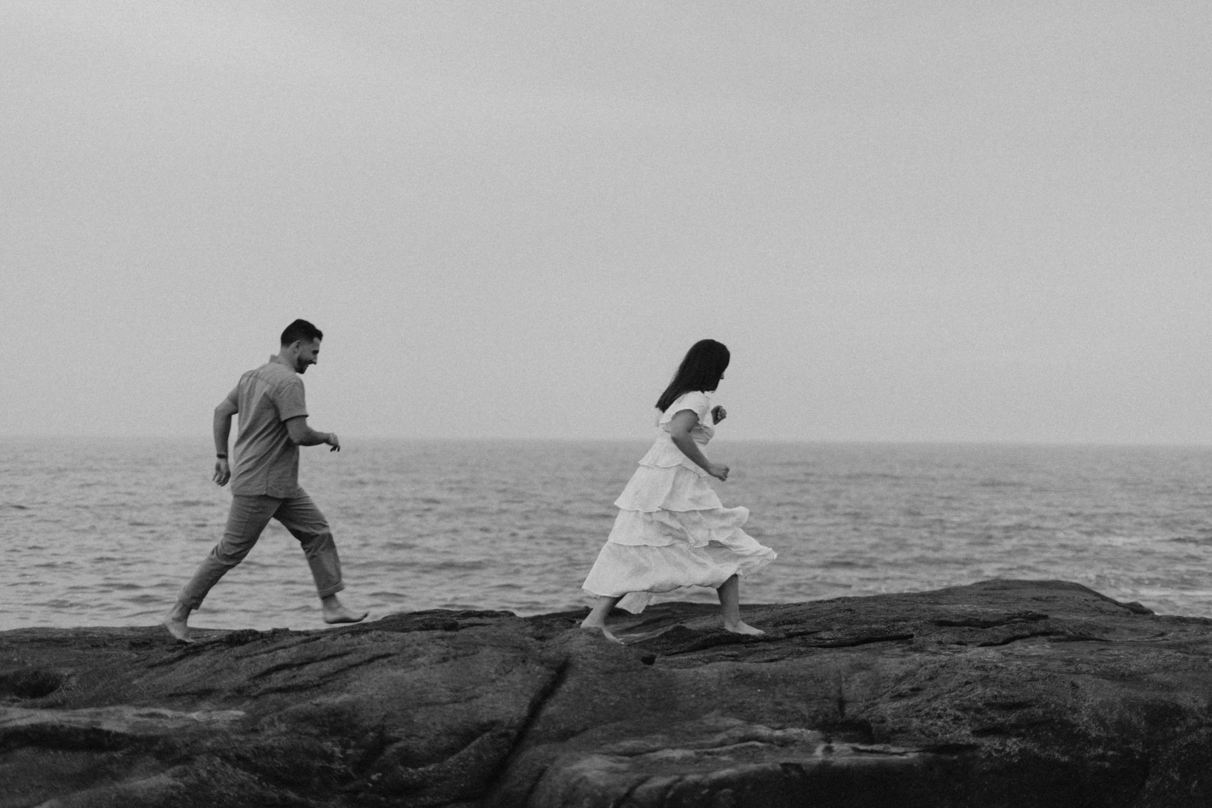 A black and white photo of a man and woman walking barefoot on a rocky shoreline, with the ocean in the background.