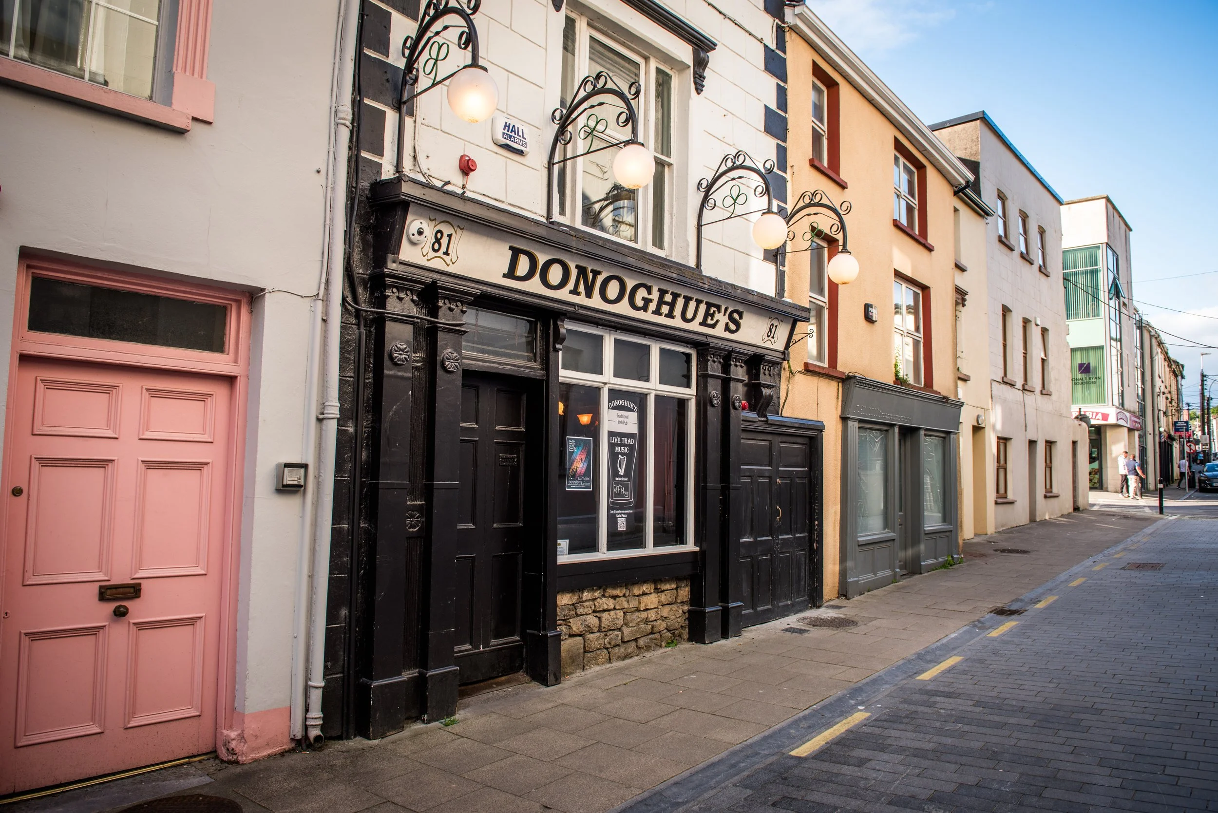 The facade of Donoghue's pub on a city street, with black framed windows and a sign reading 'DONOGHUE'S' above the entrance, and decorative spherical street lamps hanging above