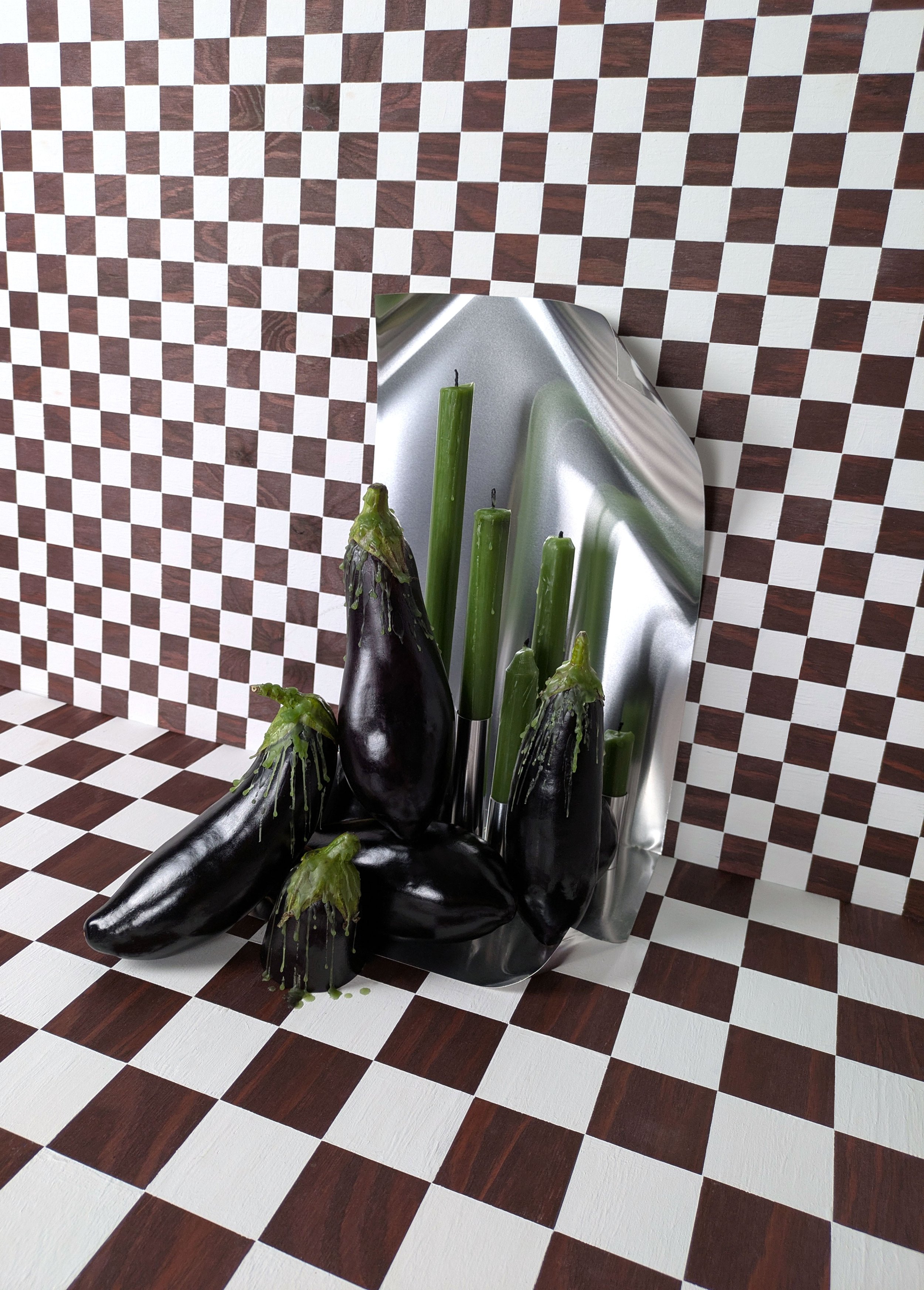 An art installation of black eggplants and green zucchini arranged against a reflective, curved backdrop on a checkered floor and wall.