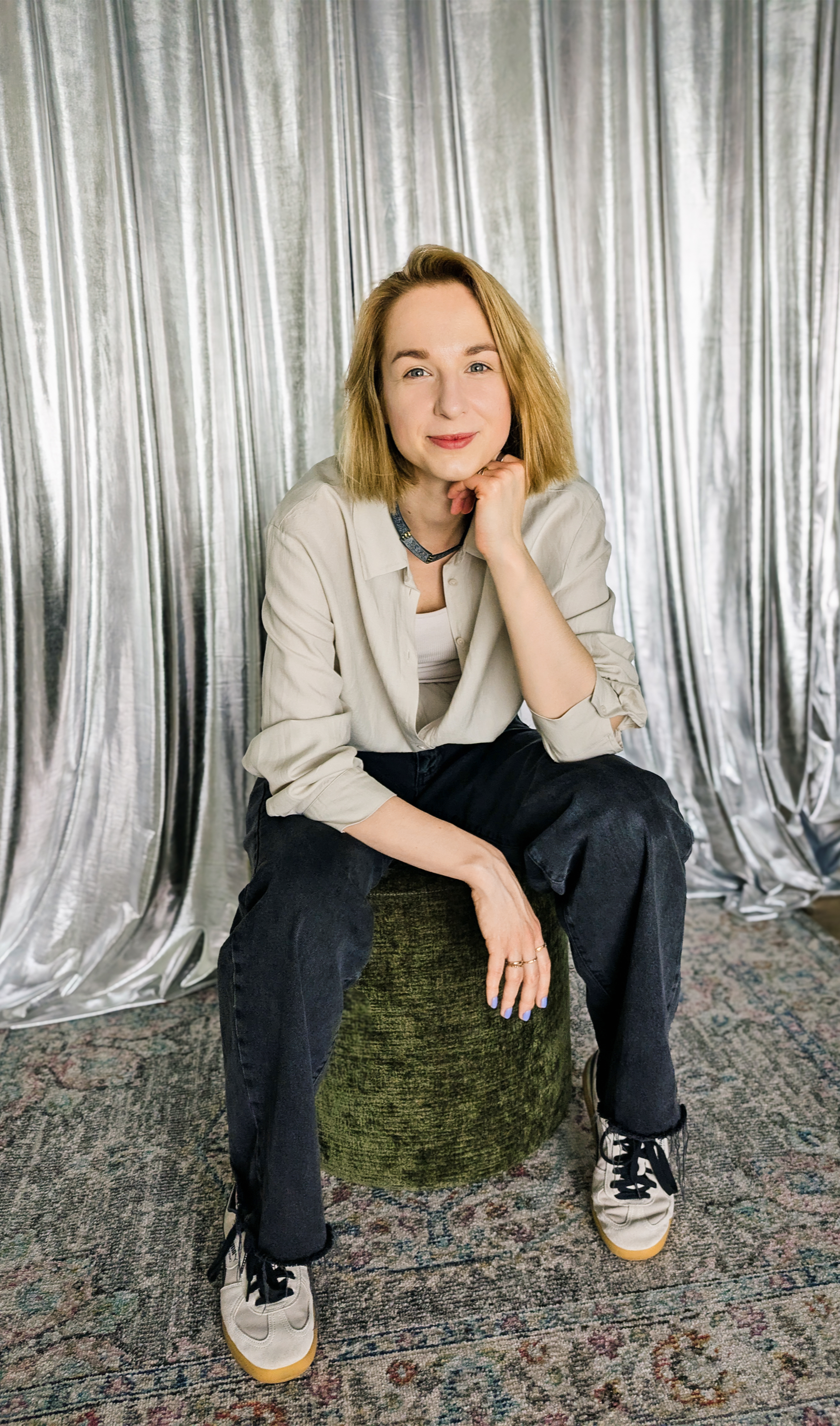 A woman with short blonde hair sitting on a green textured stool, wearing a beige shirt, dark jeans, and white sneakers, in front of a silver curtain backdrop.