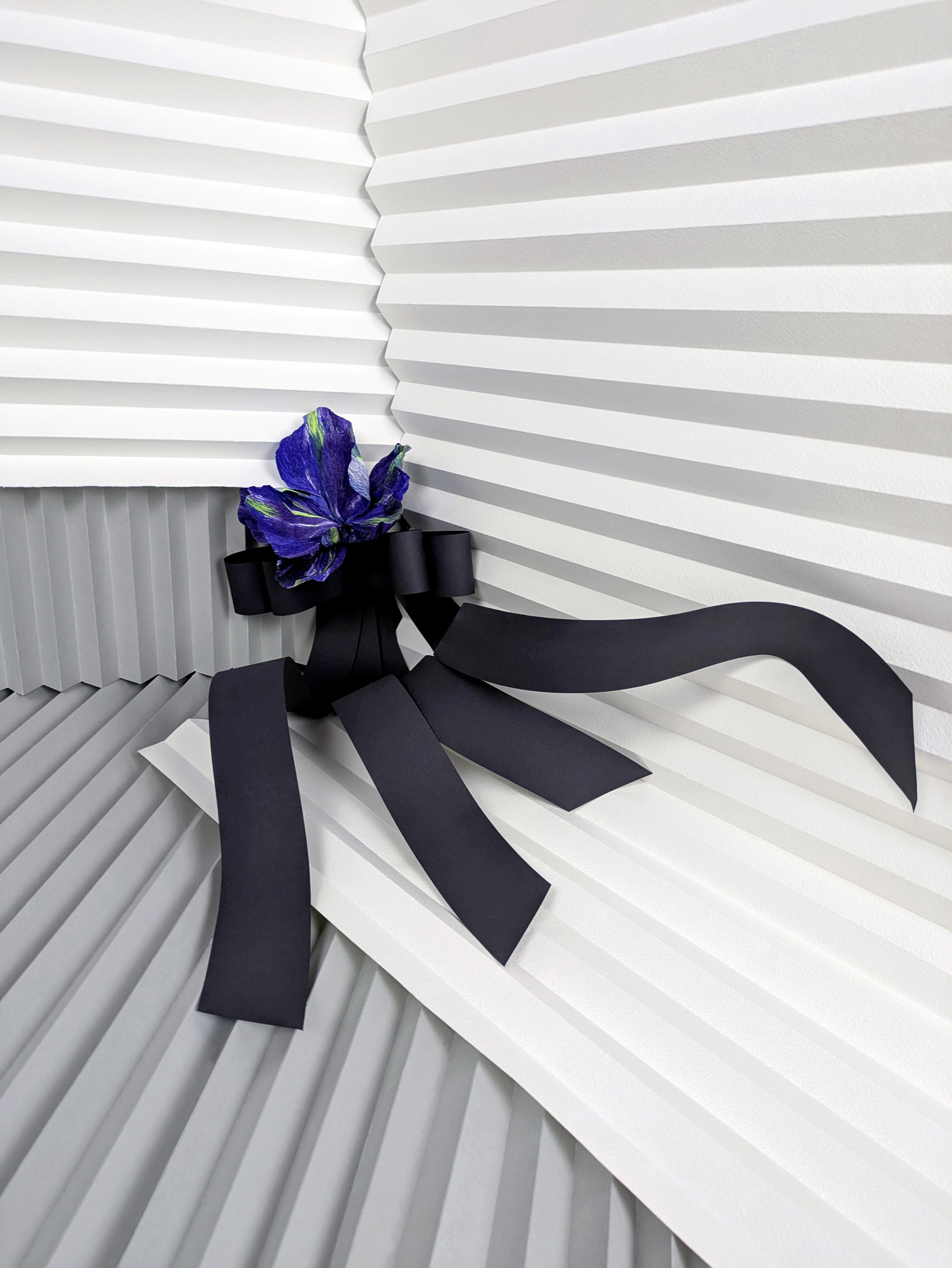 A black ribbon bow with a blue and purple flower in the center, set against a white textured background with angled ridges.