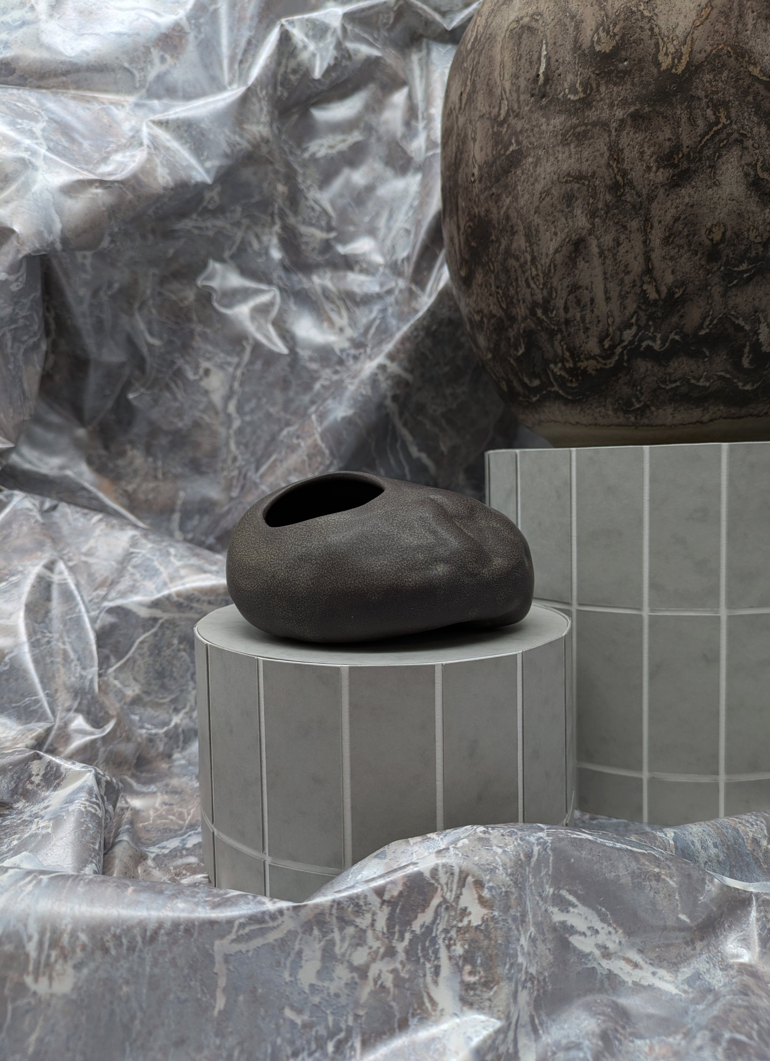 A black, smooth, rounded ceramic vase with an irregular shape and a small opening, placed on a round gray tiled pedestal, with a larger marble-like textured object and a crinkled metallic background.