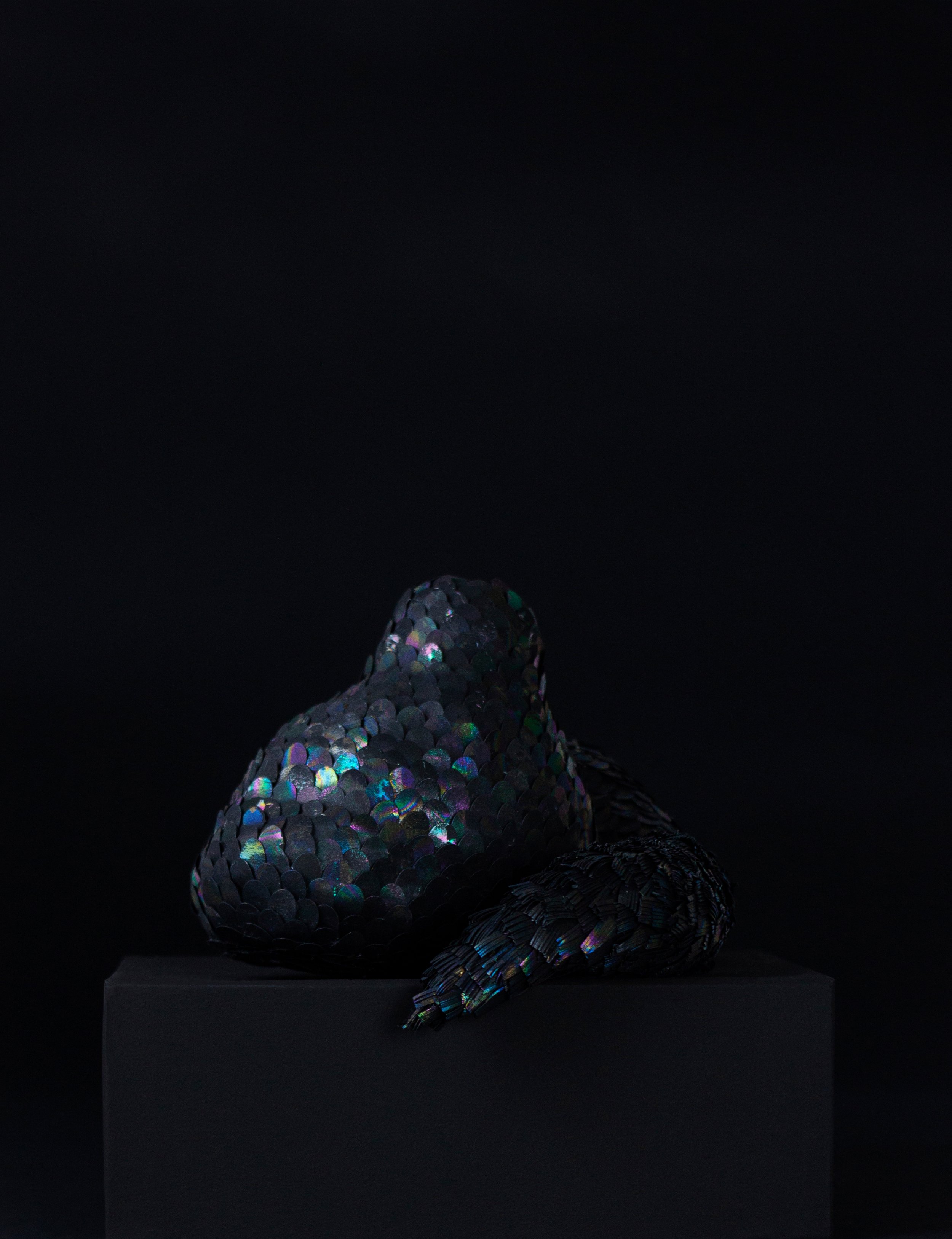 A decorative sculpture of a snake with a body covered in shiny black and multicolored iridescent scales, resting on a black pedestal against a dark background.