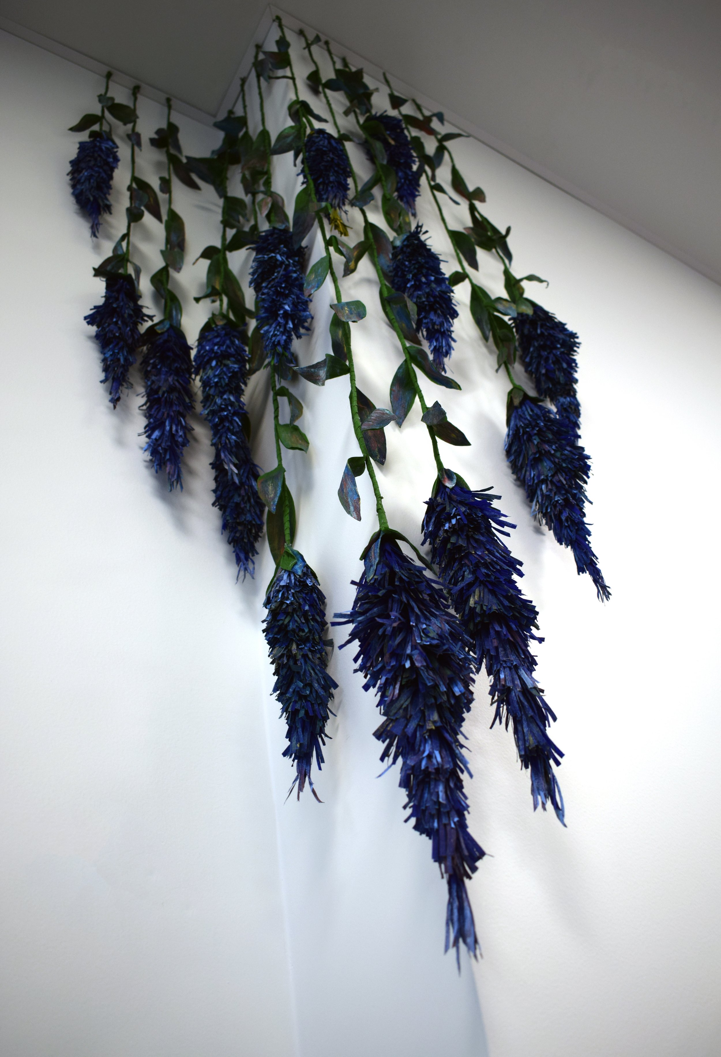 Artificial hanging flower arrangement with dark purple flowers and green leaves, attached to a white wall near the ceiling.