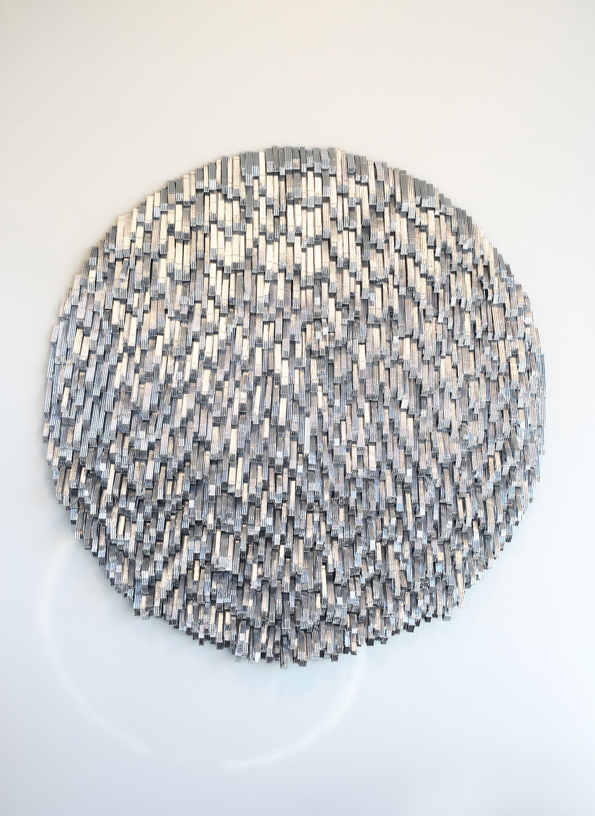A circular wall sculpture made of numerous metallic rectangular pieces arranged in a textured, layered pattern.