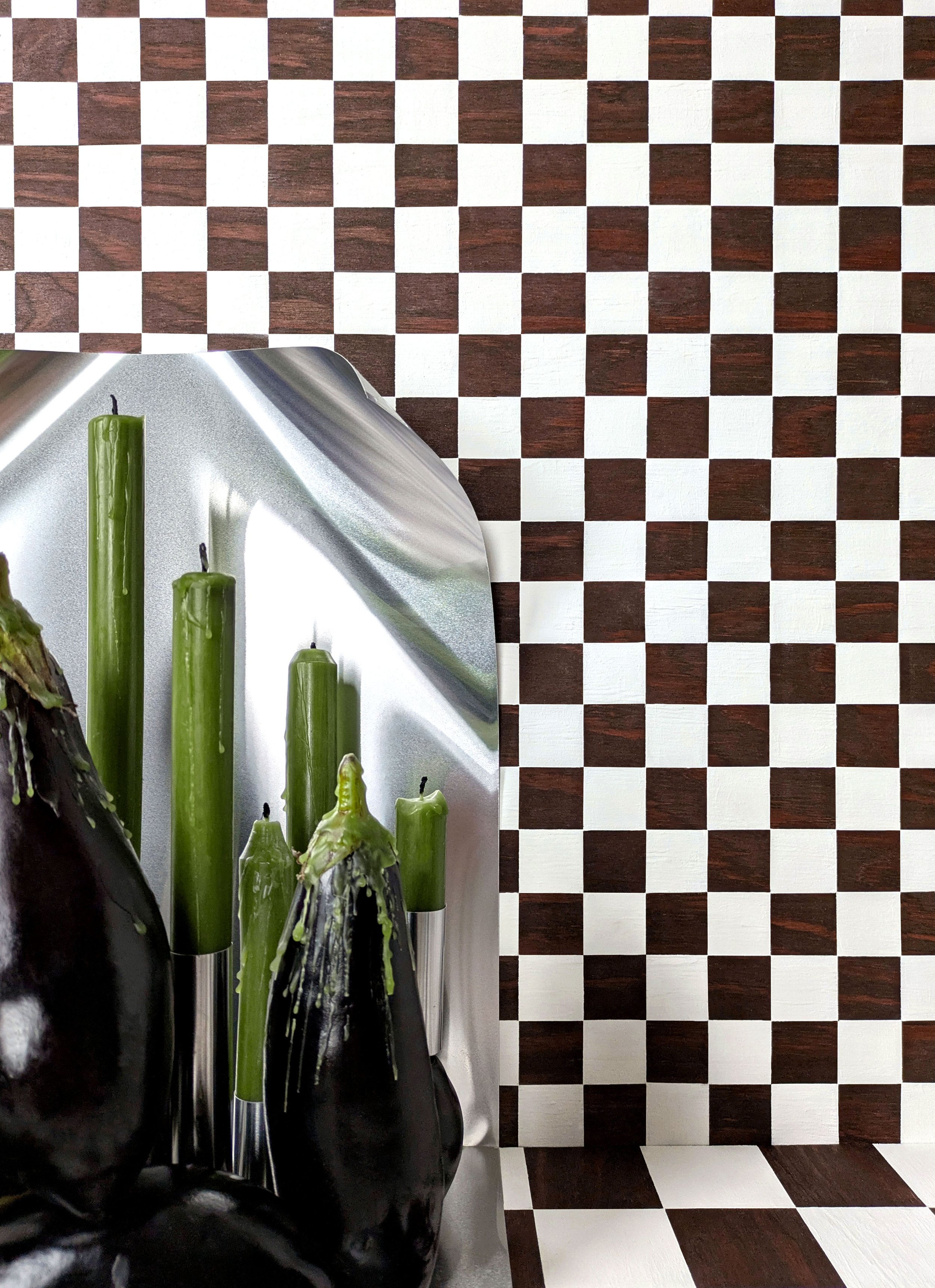 A geometric interior design with a checkered pattern on the wall and floor, featuring green candles and black eggplants with blemishes.