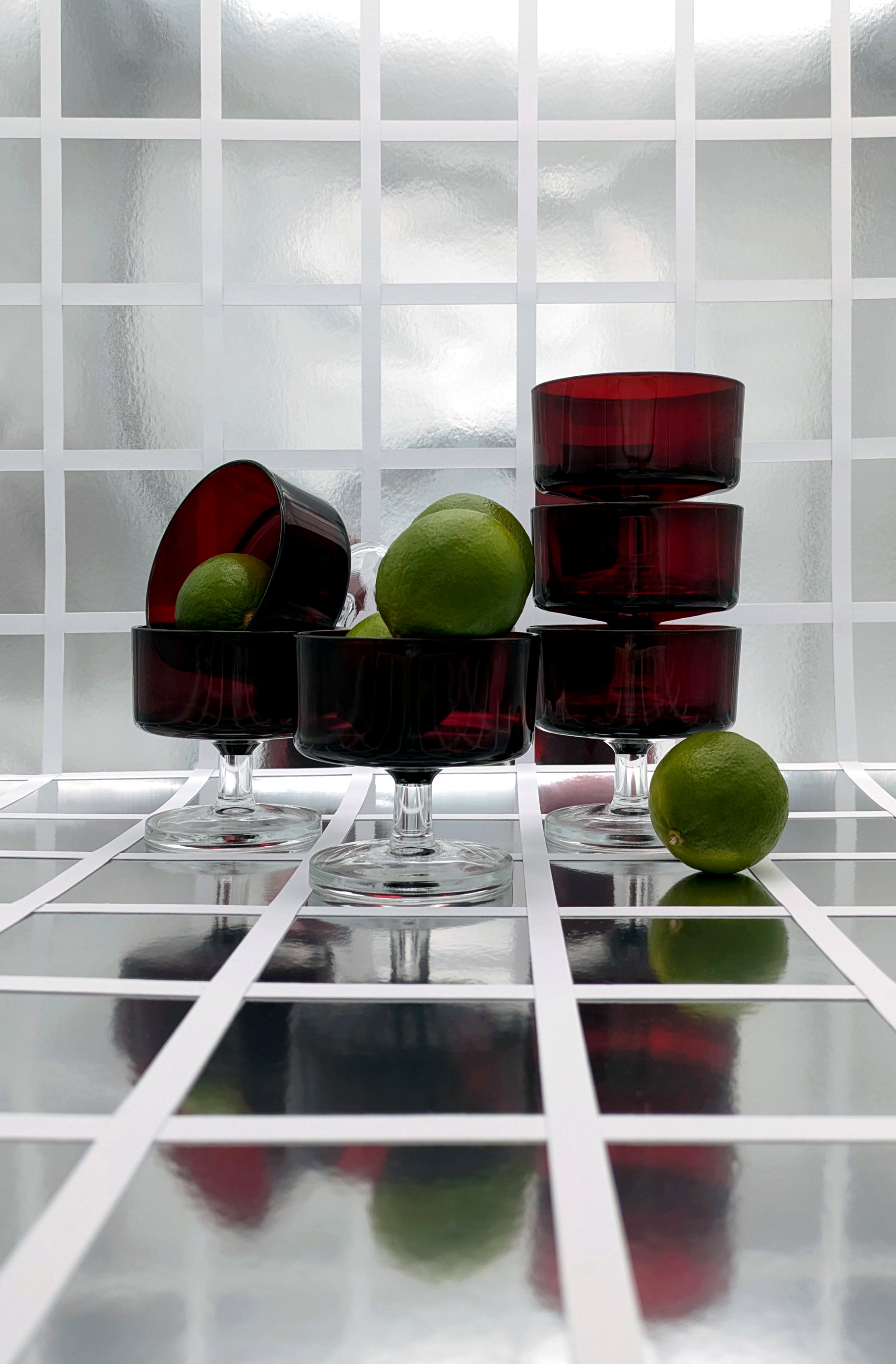 Several red glass bowls stacked and one lying on its side, with green limes inside and around, set against a frosted glass block background and on a reflective grid surface.