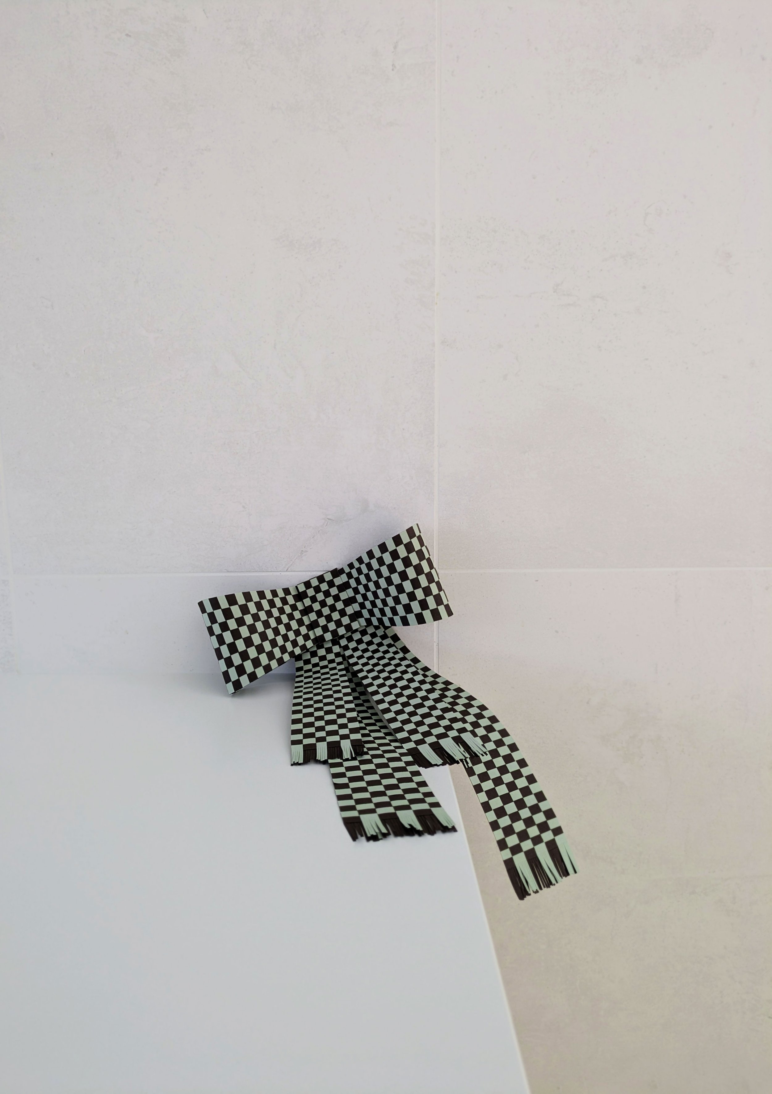A black and white checkered bowtie and scarf set placed on a white surface against a textured white wall background.