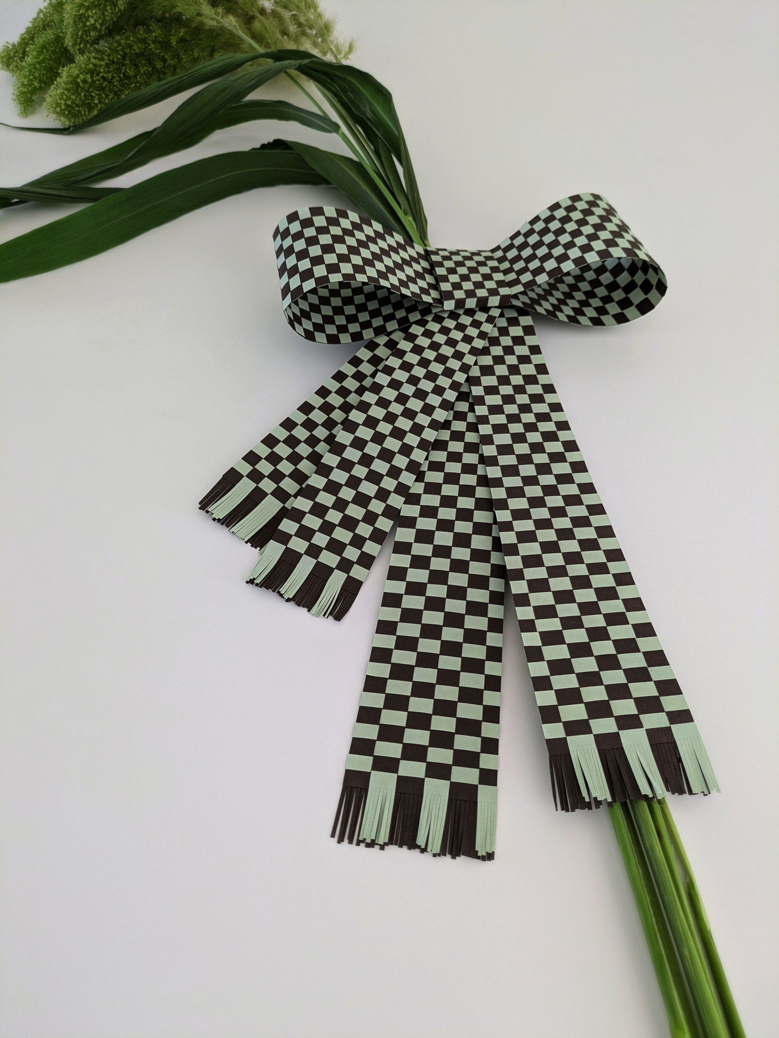 A checkered black and white ribbon tied in a bow, attached to green flowers and leaves on a white background.