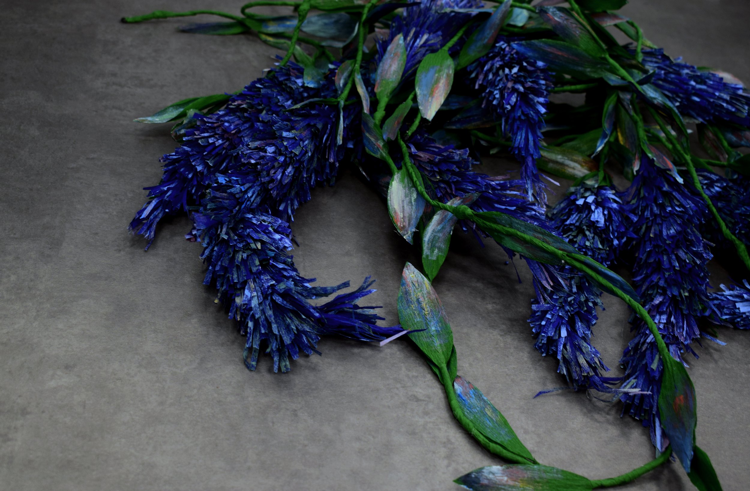 A bunch of artificial purple flowers with green leaves, laying on a gray surface.