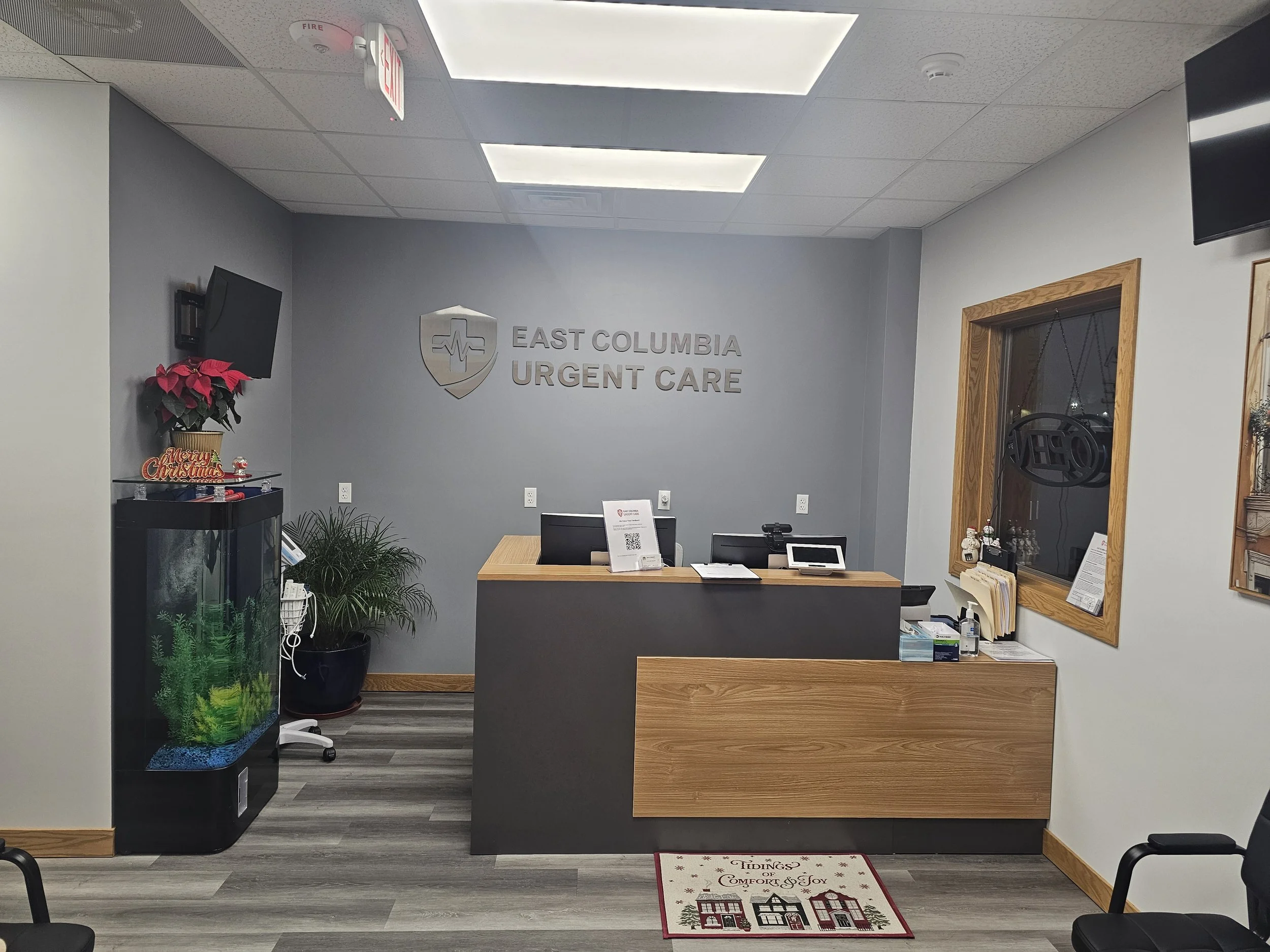 The reception area of East Columbia Urgent Care with a desk, two computer monitors, a QR code sign, a poinsettia plant, a fish tank, and holiday decorations including a "Merry Christmas" sign and a small gift box with a wreath hanging on the window.