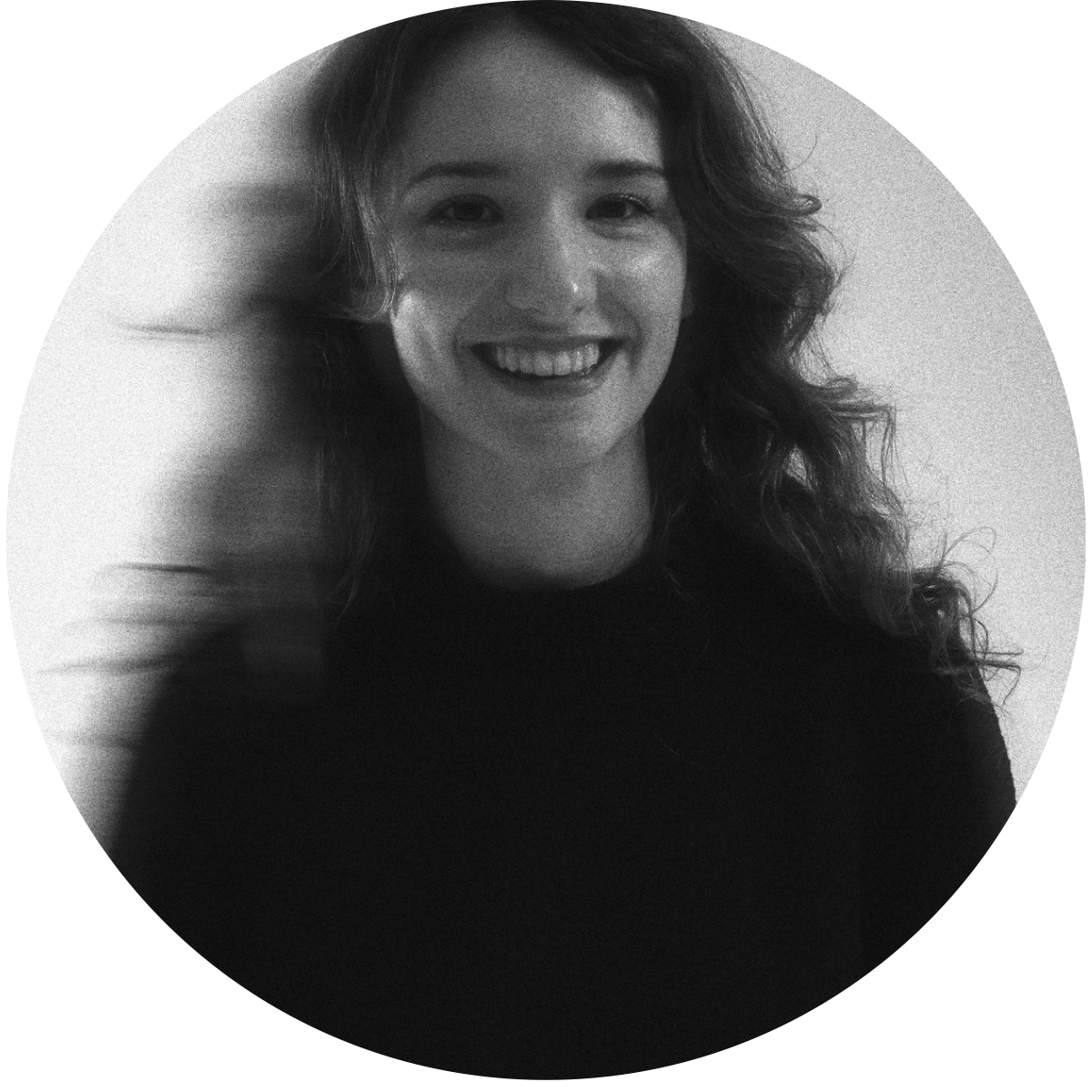Black and white photo of a woman smiling with curly hair, wearing a dark top, with one hand touching her head