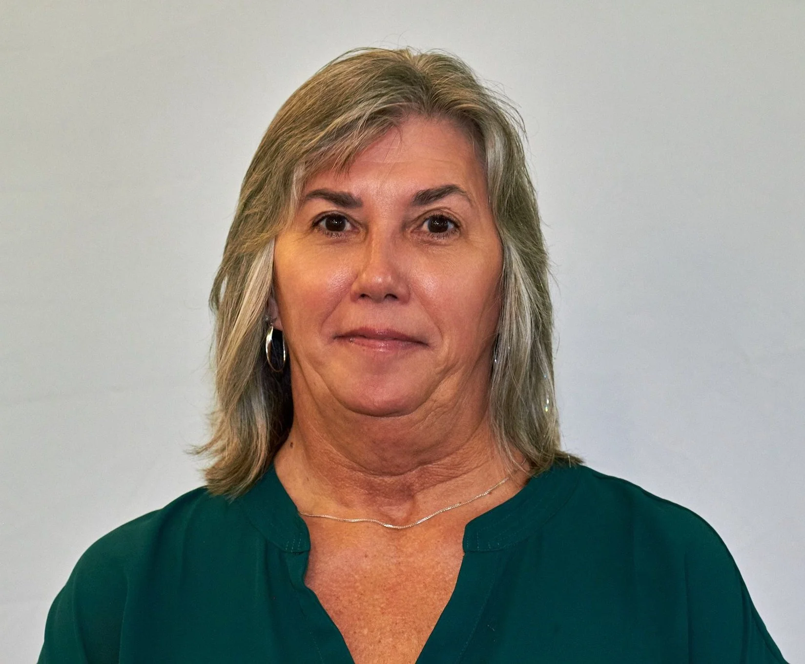 A woman with gray hair wearing a green patterned blouse smiles at the camera against a plain background. She is wearing earrings and a necklace.